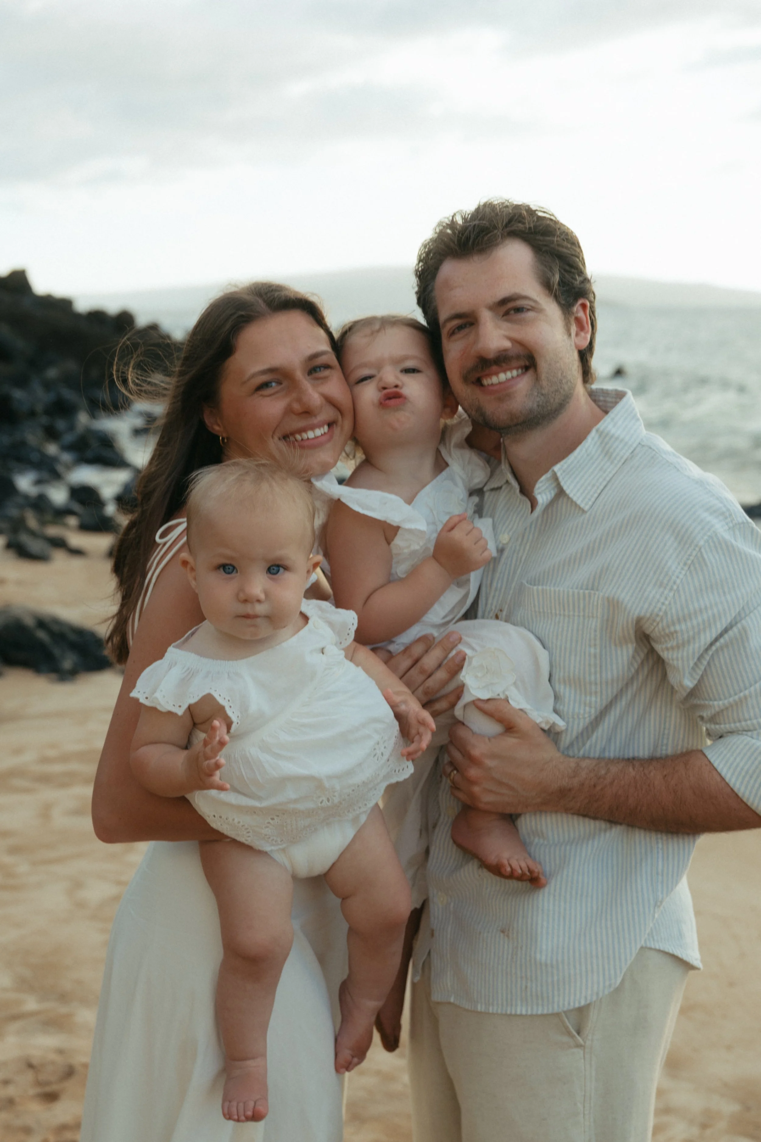 A happy family of four, including a woman, a man, and two young children, posing together on a beach with sand and rocks, with the ocean and cloudy sky in the background.