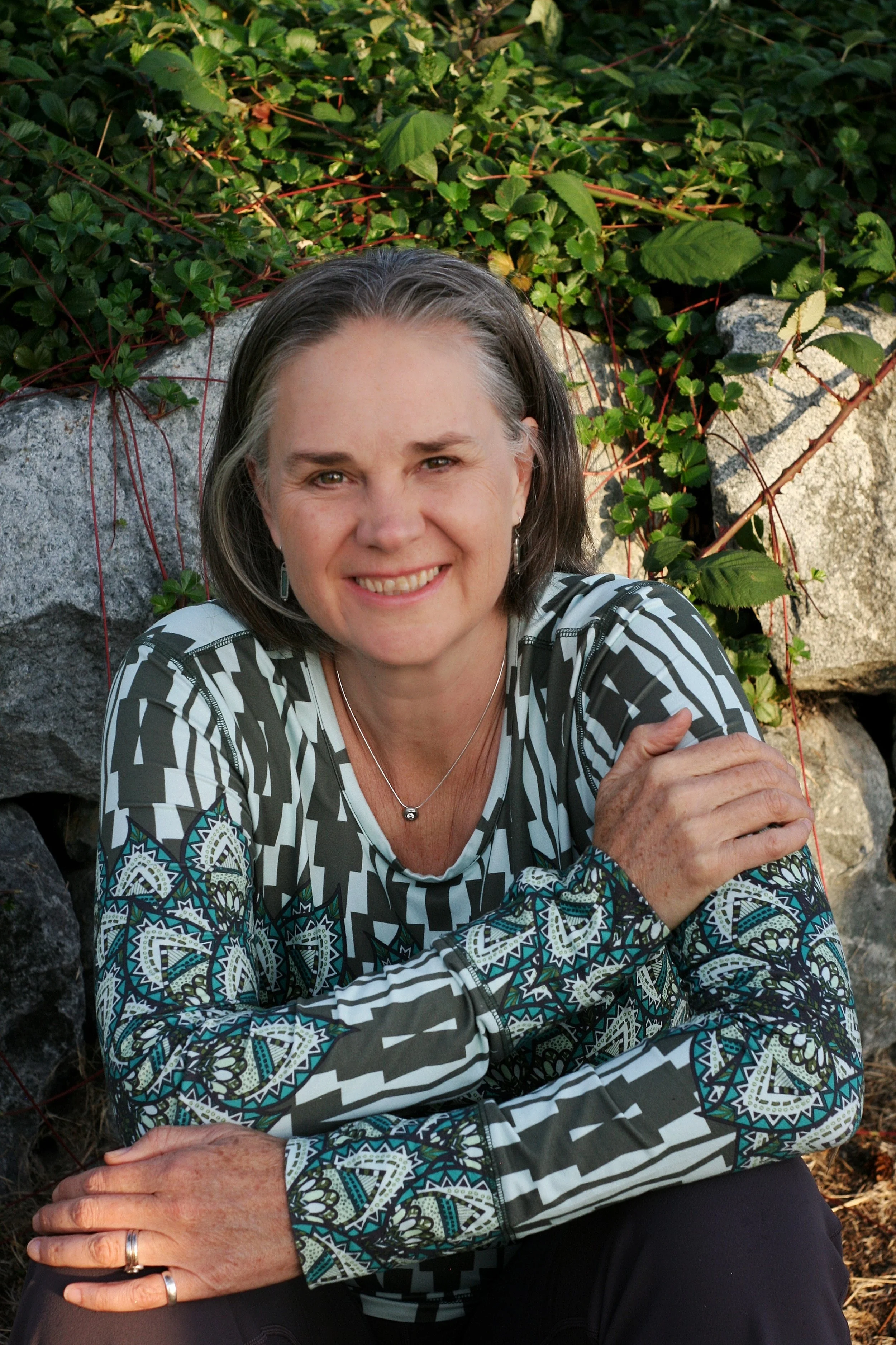 A woman with shoulder-length gray hair sitting outdoors in front of a stone wall with green plants, smiling at the camera.
