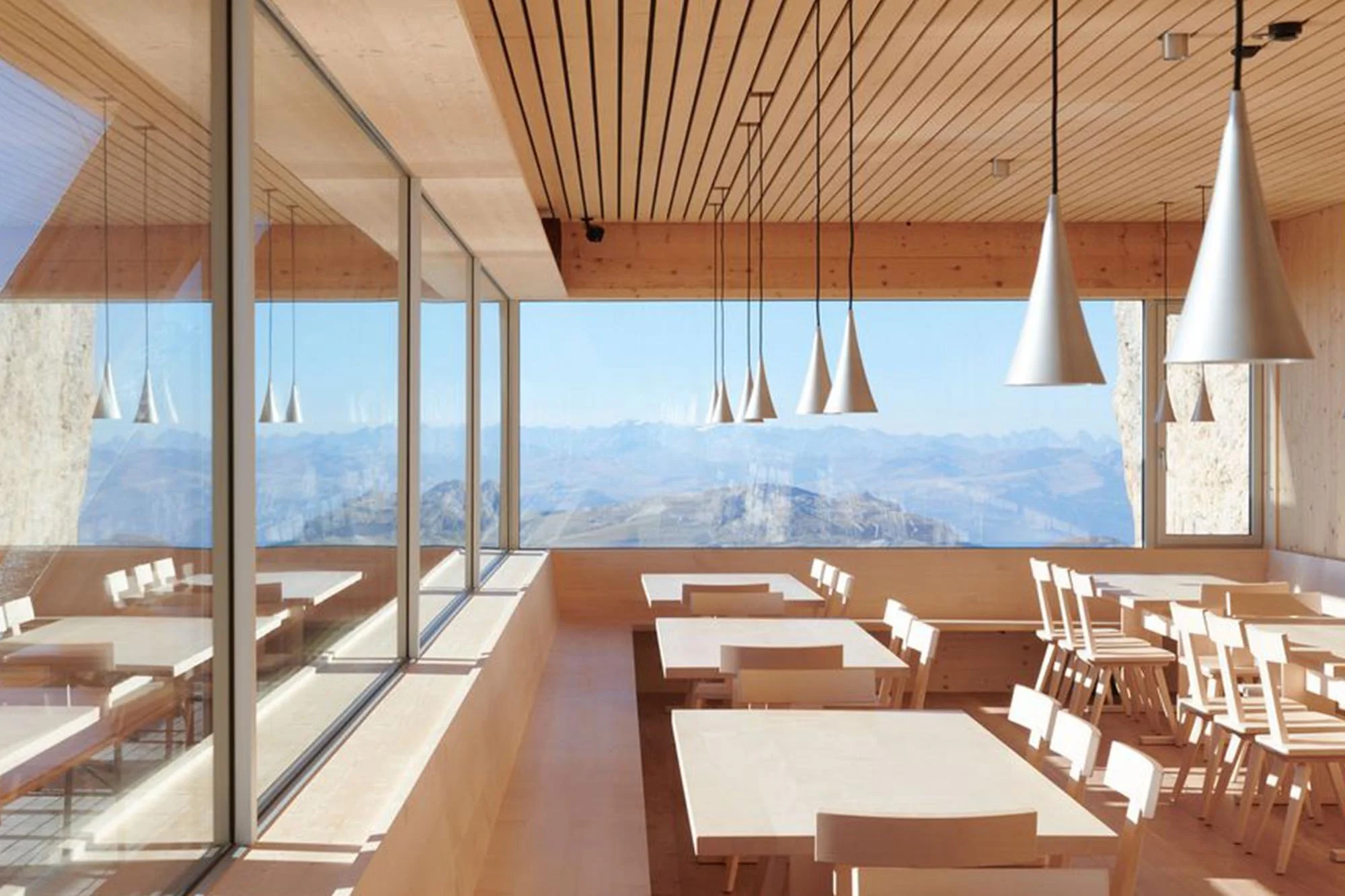 Dining room inside the Santnerpass Hut with timber ceilings, pendant lights and panoramic windows overlooking the Dolomites.