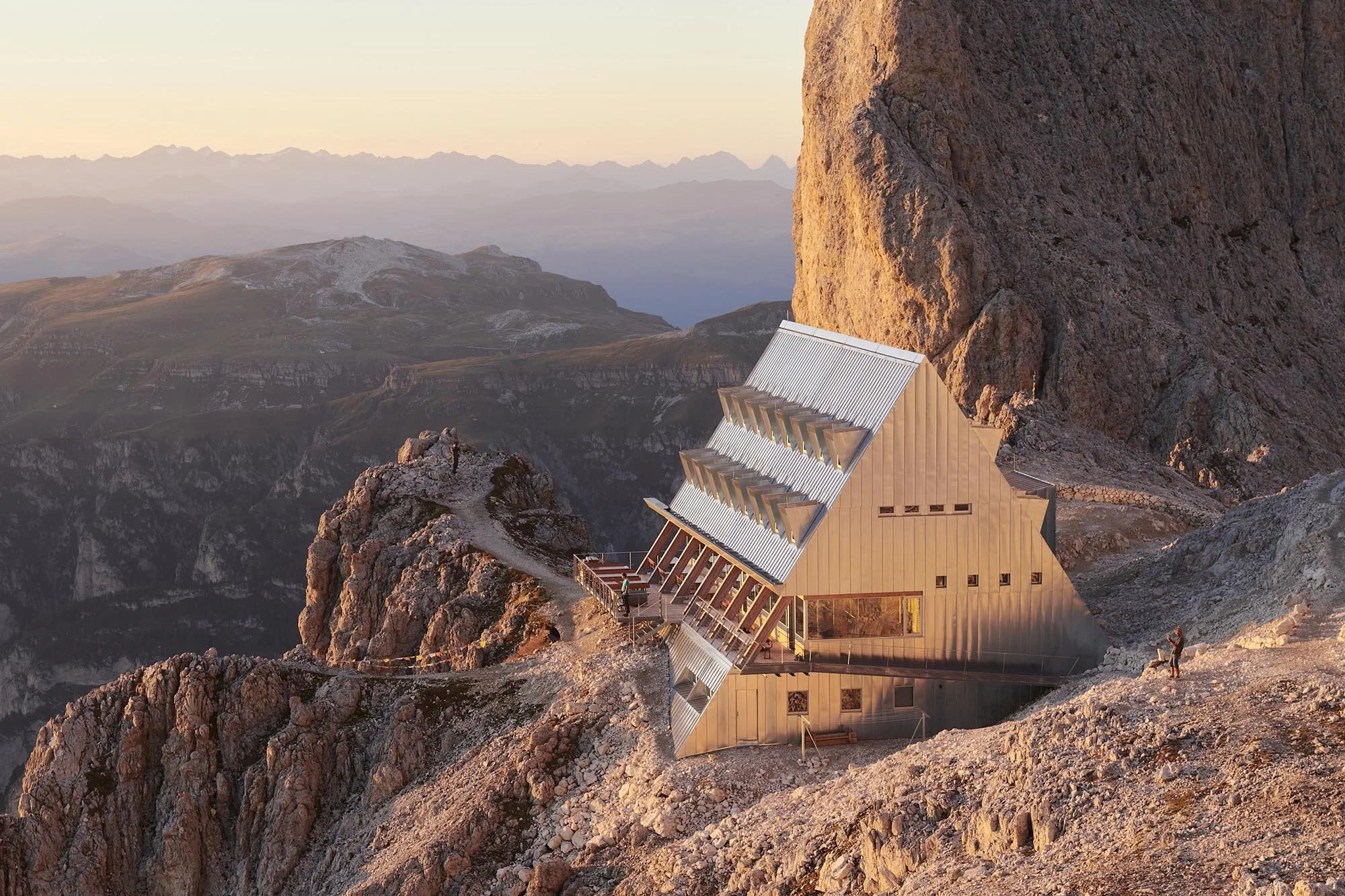 Santnerpass Hut in warm evening light, showing its steep galvanised-steel roof against the Rosengarten rock face.
