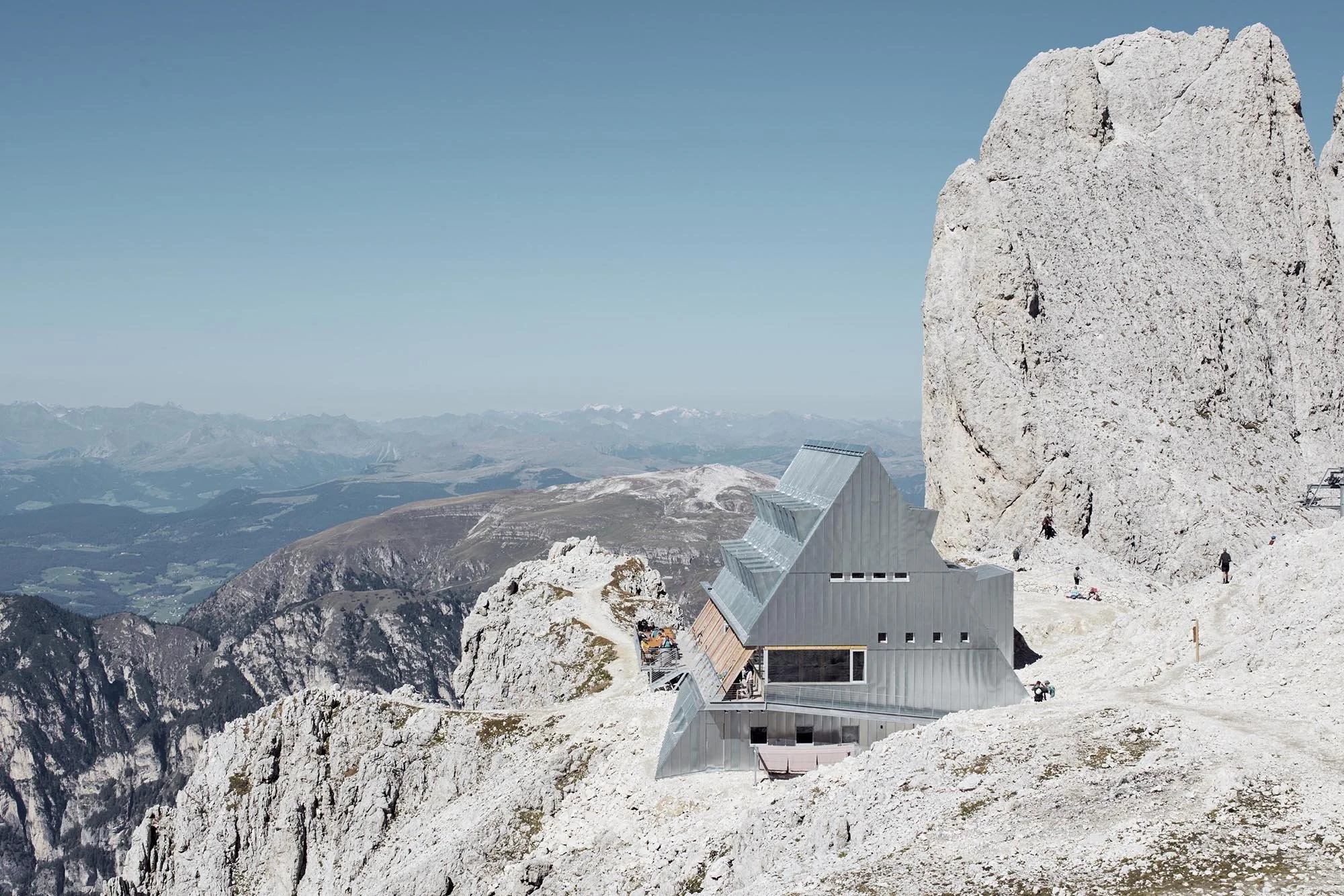 Santnerpass Hut in warm evening light, showing its steep galvanised-steel roof against the Rosengarten rock face.