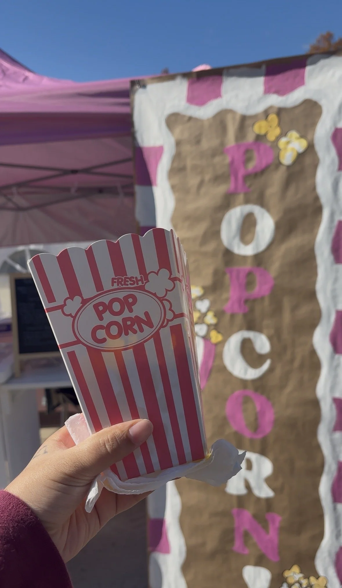 Image of a pink popcorn container next to a painted vertical sign that says 'popcorn' in alternating pink and white colors.