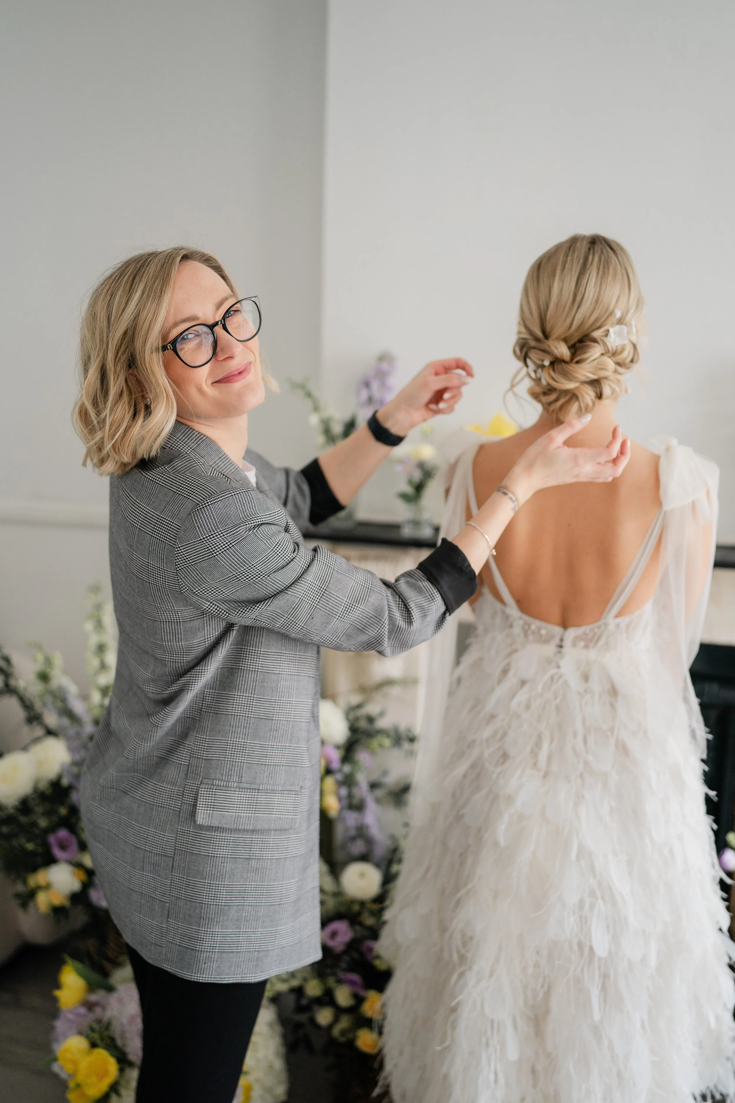 A hairstylist works on a bride's updo hairstyle while the bride wears a white dress with feather details. Flowers are arranged in the background.
