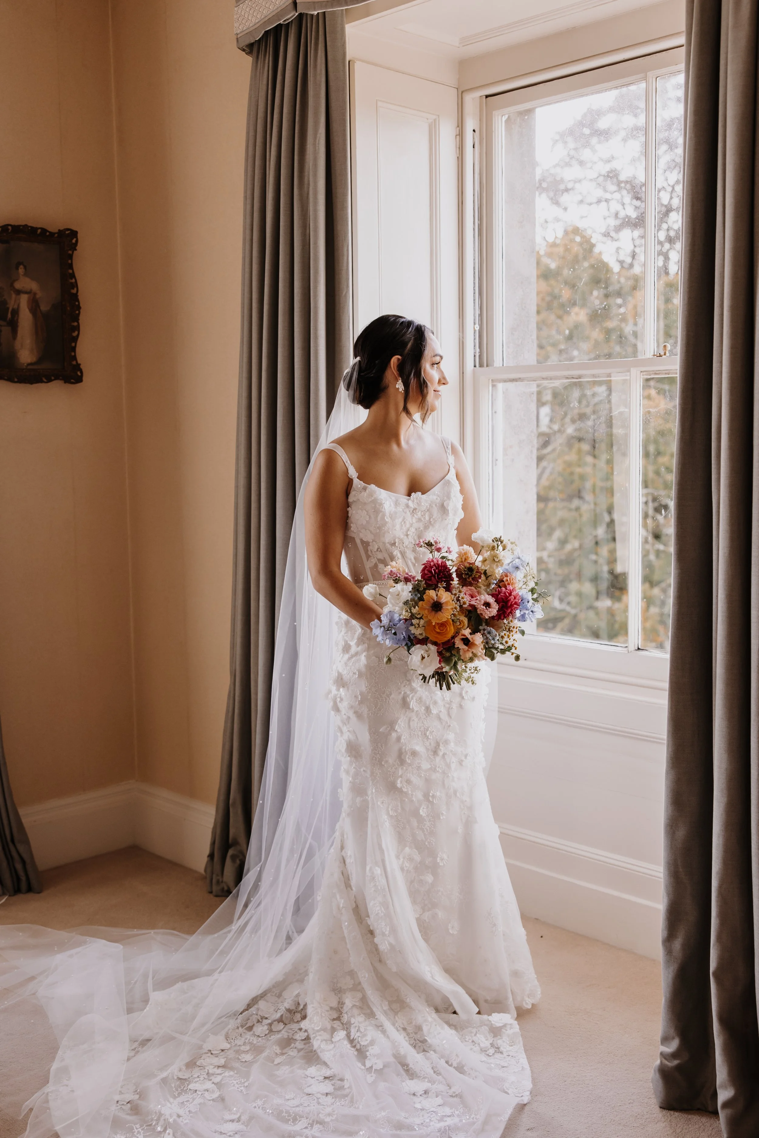 Bride in lace wedding dress holding bouquet by window at Somerley House, Ringwood, Cotswolds, Wiltshire wedding florist.