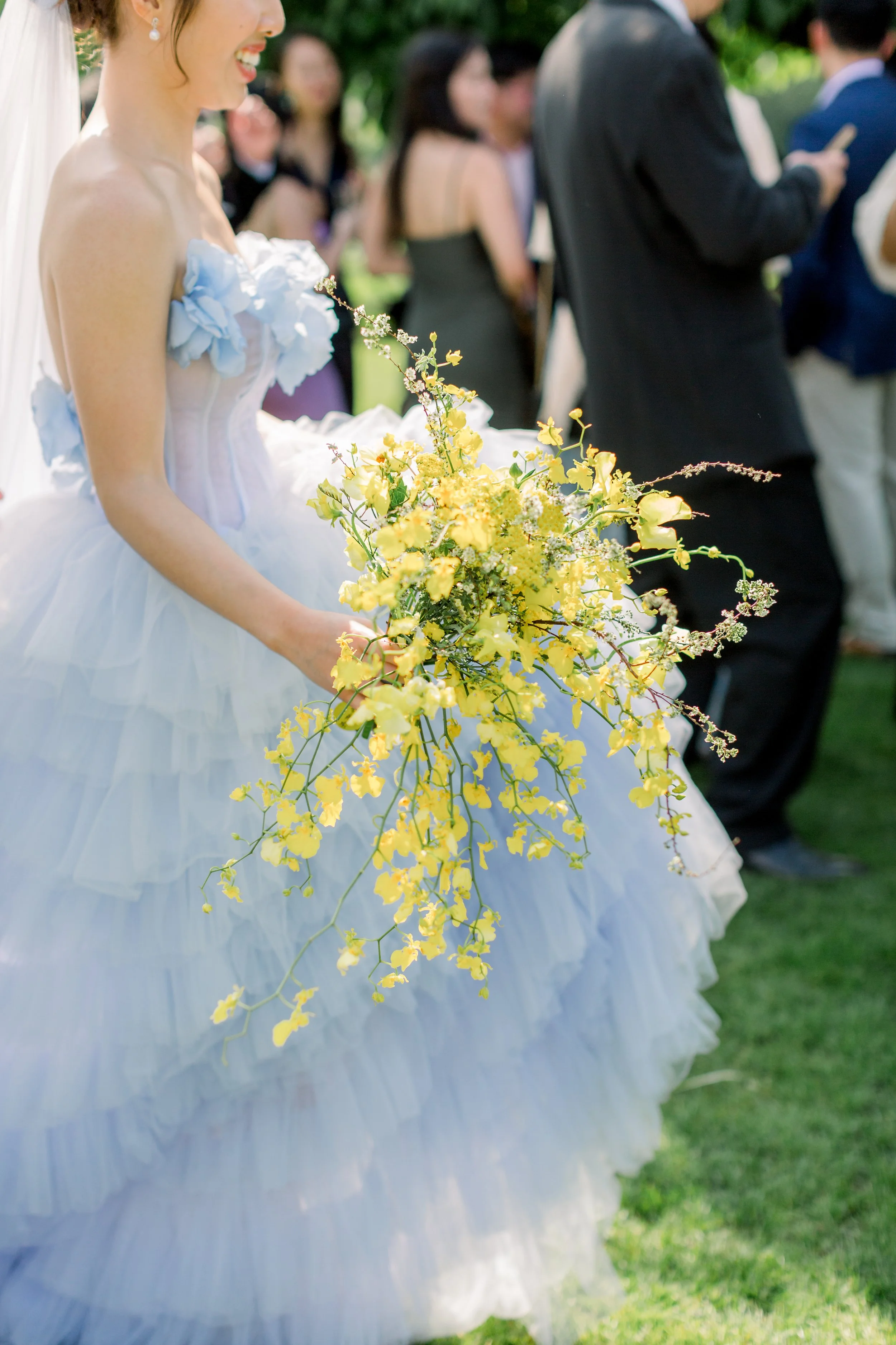 Bride in light blue ruffled dress holding a floral umbrella, standing in a garden decorated with greenery and colorful flowers at Hyde House, Cotswolds florist. 