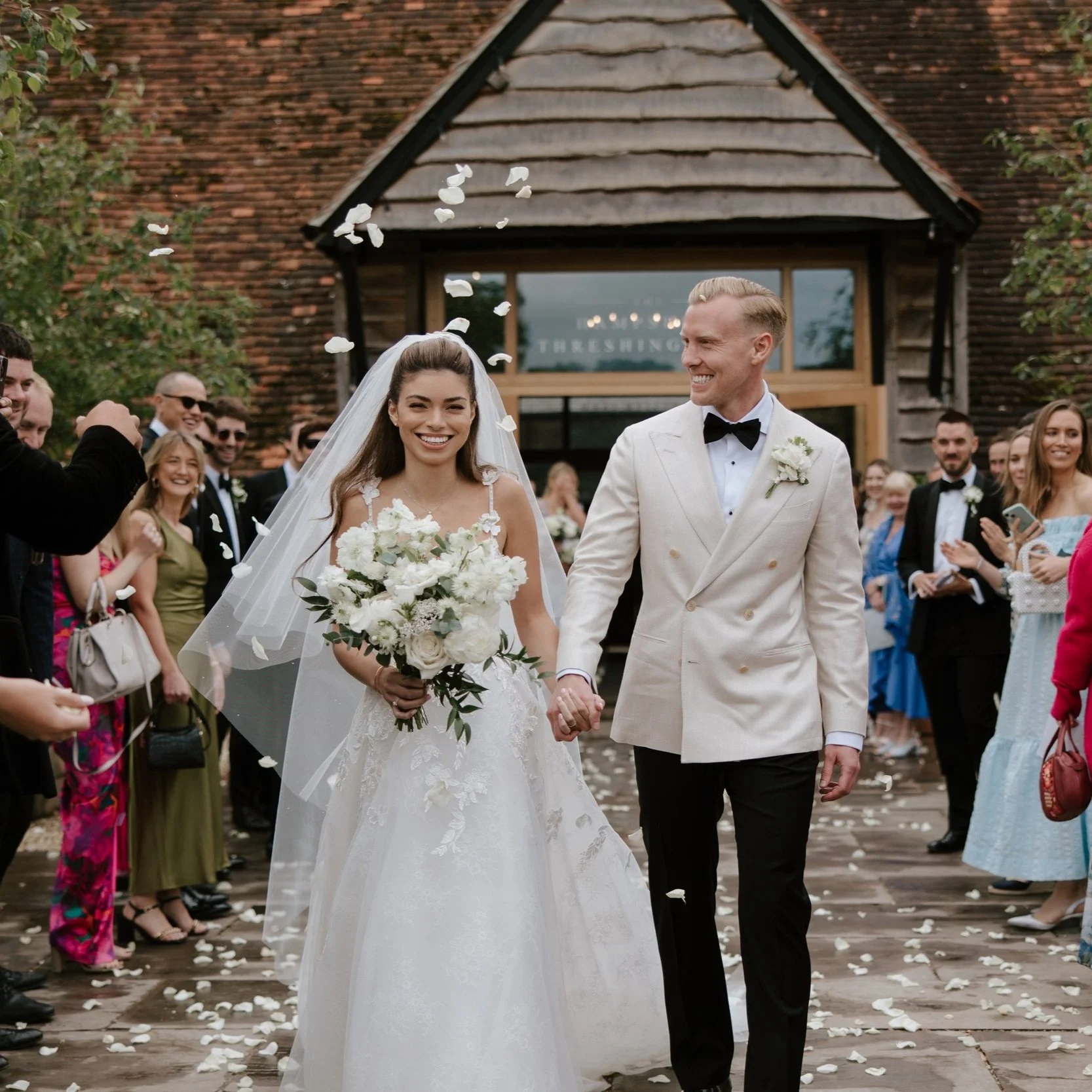 Bride and groom walking down the aisle with guests celebrating, bride holding a bouquet, groom in a cream double-breasted suit at a Cotswold wedding florist.