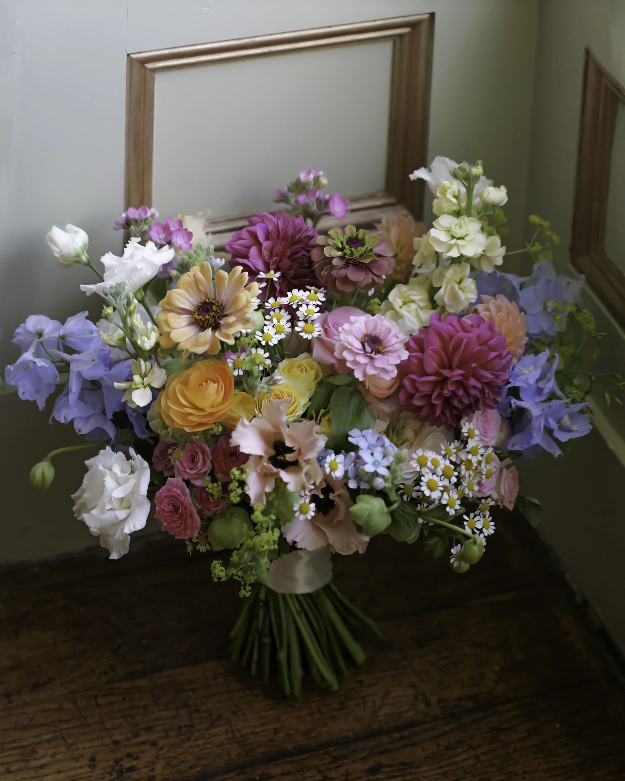 Colorful floral bouquet in a rustic setting with mixed flowers including roses, daisies, and dahlias in shades of pink, purple, yellow, and white.
