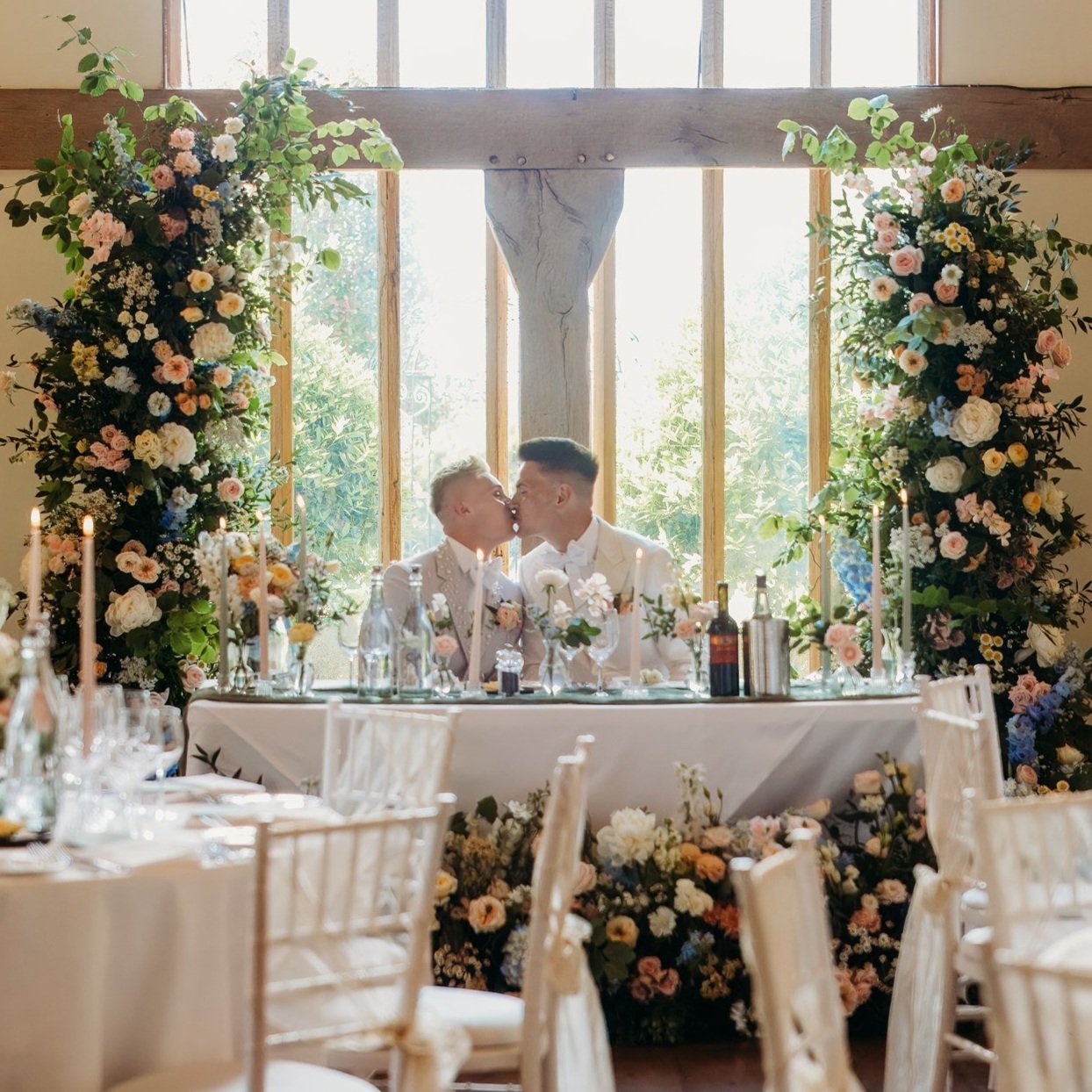 Two people kissing at a wedding reception, seated at a decorated table with floral arrangements and candles, in front of large windows. Oxfordshire wedding florist.