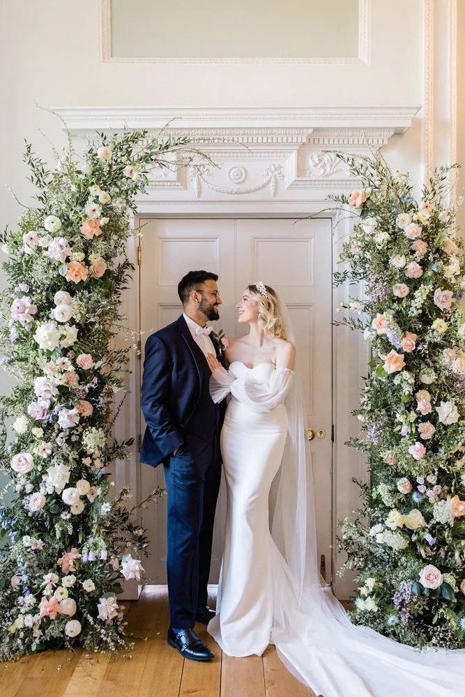A couple standing in front of a decorated doorway, surrounded by floral arrangements. The bride is wearing a white gown and veil, while the groom is in a dark suit. They are smiling at each other.