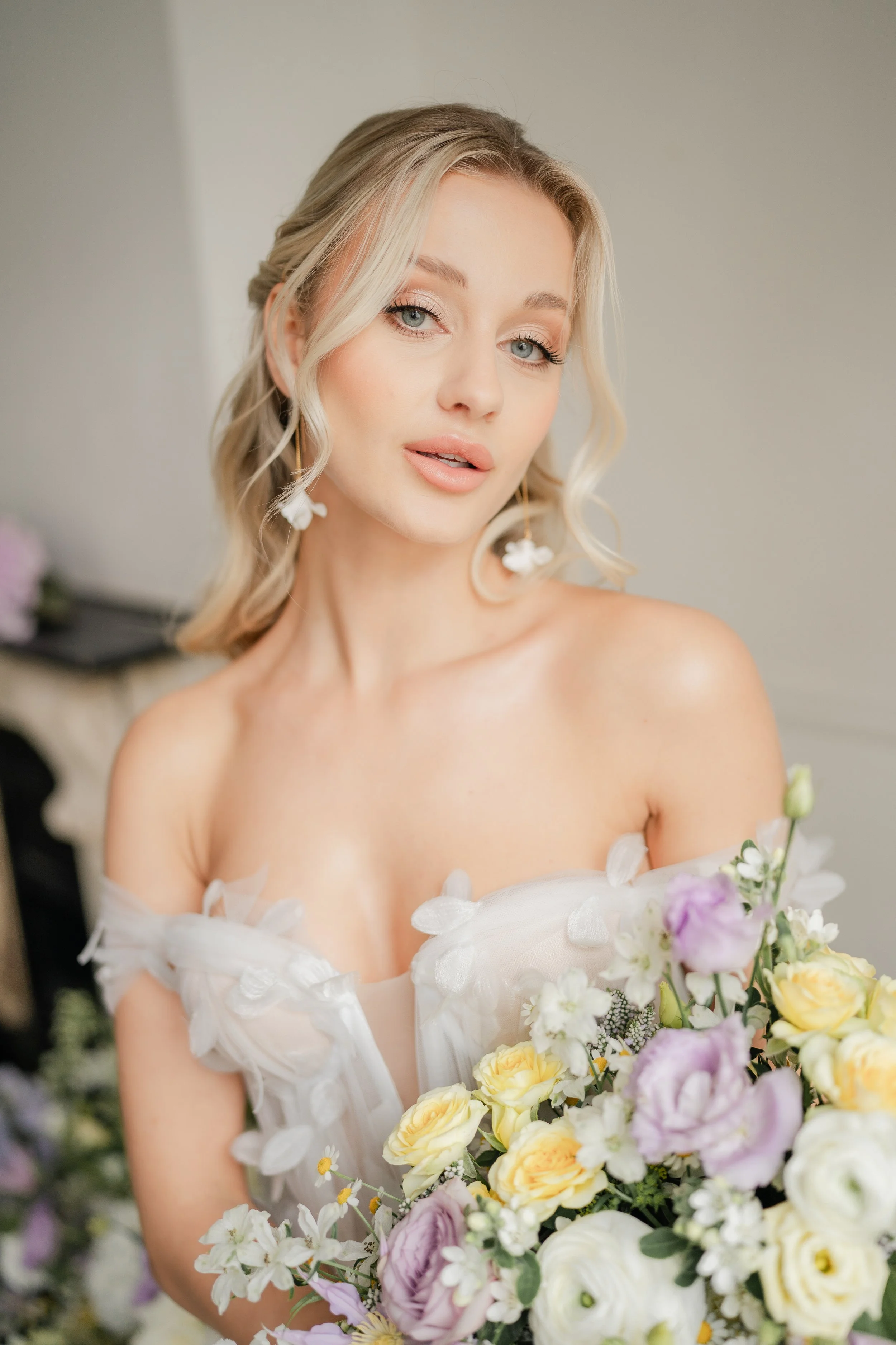 Woman with blonde hair holding a bouquet of yellow and purple flowers, wearing a white off-the-shoulder dress with floral details.