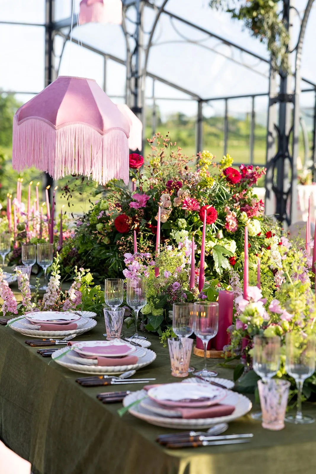 Elegant wedding reception setup with long tables, white tablecloths, floral centerpieces, candles, and green cushioned chairs in an airy venue with white columns and greenery at Avington Park. 