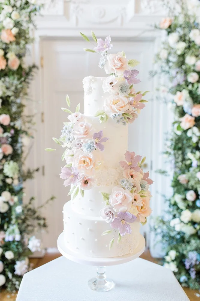 Elegant three-tier white wedding cake adorned with pastel-colored floral decorations, displayed on a glass stand with floral arrangements in the background.