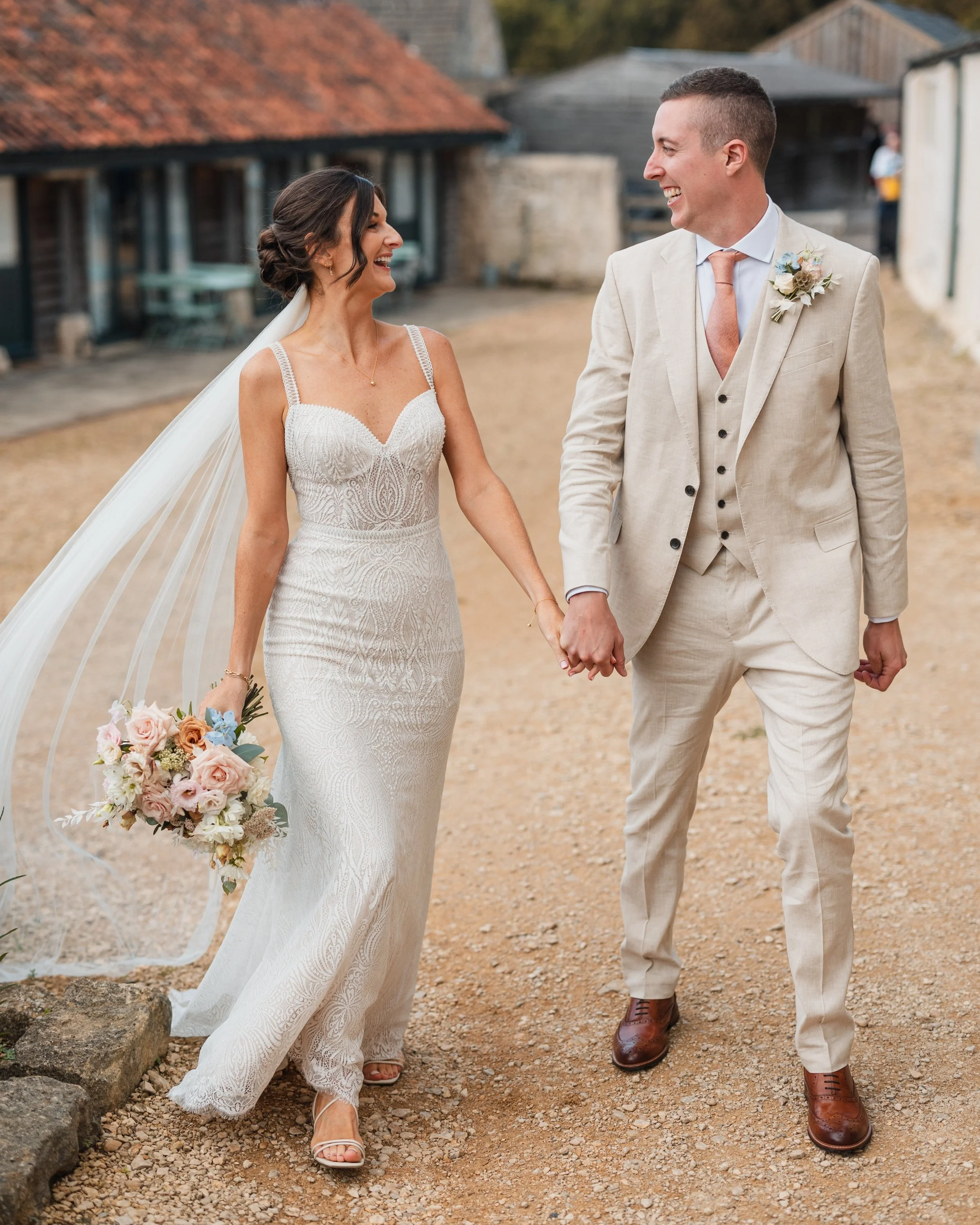 Bride and groom holding hands, bride in lace wedding dress holding bouquet, groom in beige suit, outdoor setting at a Bath wedding venue, florist.