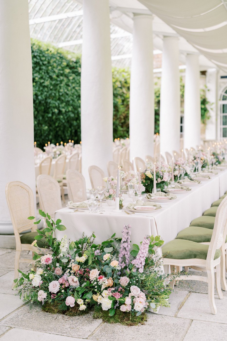 Elegant wedding reception setup with long tables, white tablecloths, floral centerpieces, candles, and green cushioned chairs in an airy venue with white columns and greenery at Avington Park.