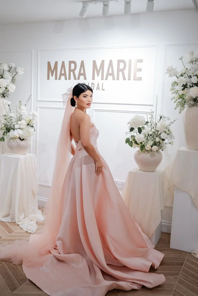 Woman in a pink bridal gown posing in front of a "Mara Marie Bridal" sign, surrounded by white floral arrangements.