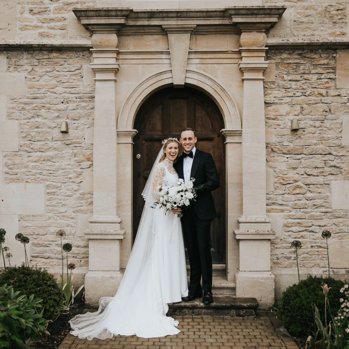 A bride and groom standing together in front of a stone building, the bride wearing a white gown and holding a bouquet, the groom in a black tuxedo at Kin House, Kin House Wiltshire, Kin House Florist.
