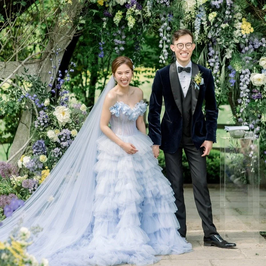 Wedding couple standing at an outdoor ceremony with floral arch, bride in blue tiered gown, groom in dark tuxedo at Hyde House, Cotswolds Florist