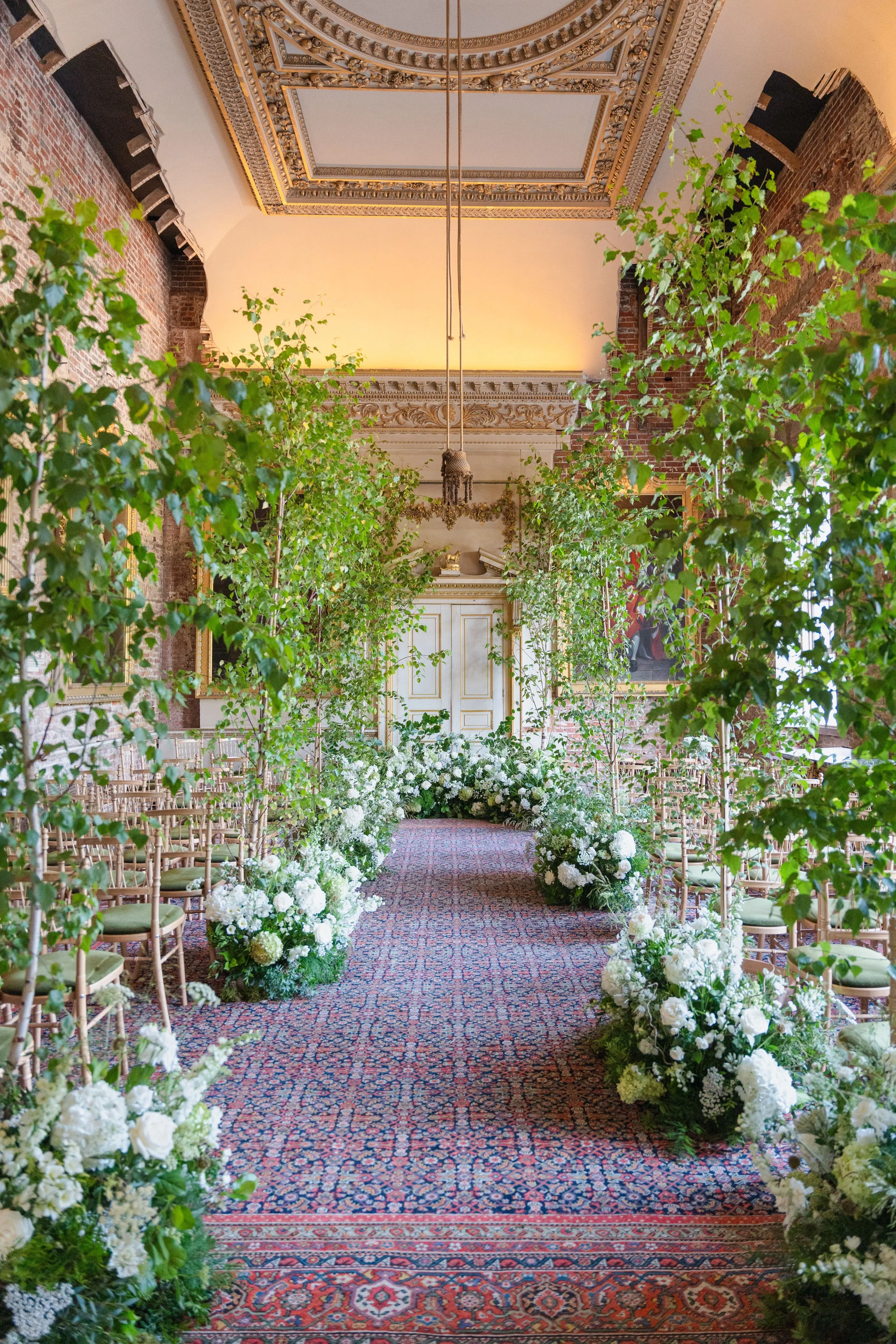 Elegant wedding aisle decorated with white flowers and greenery, lined with gold chairs in an ornate room with a decorative ceiling and large carpet at St. Giles House Wedding, florist Dorset.