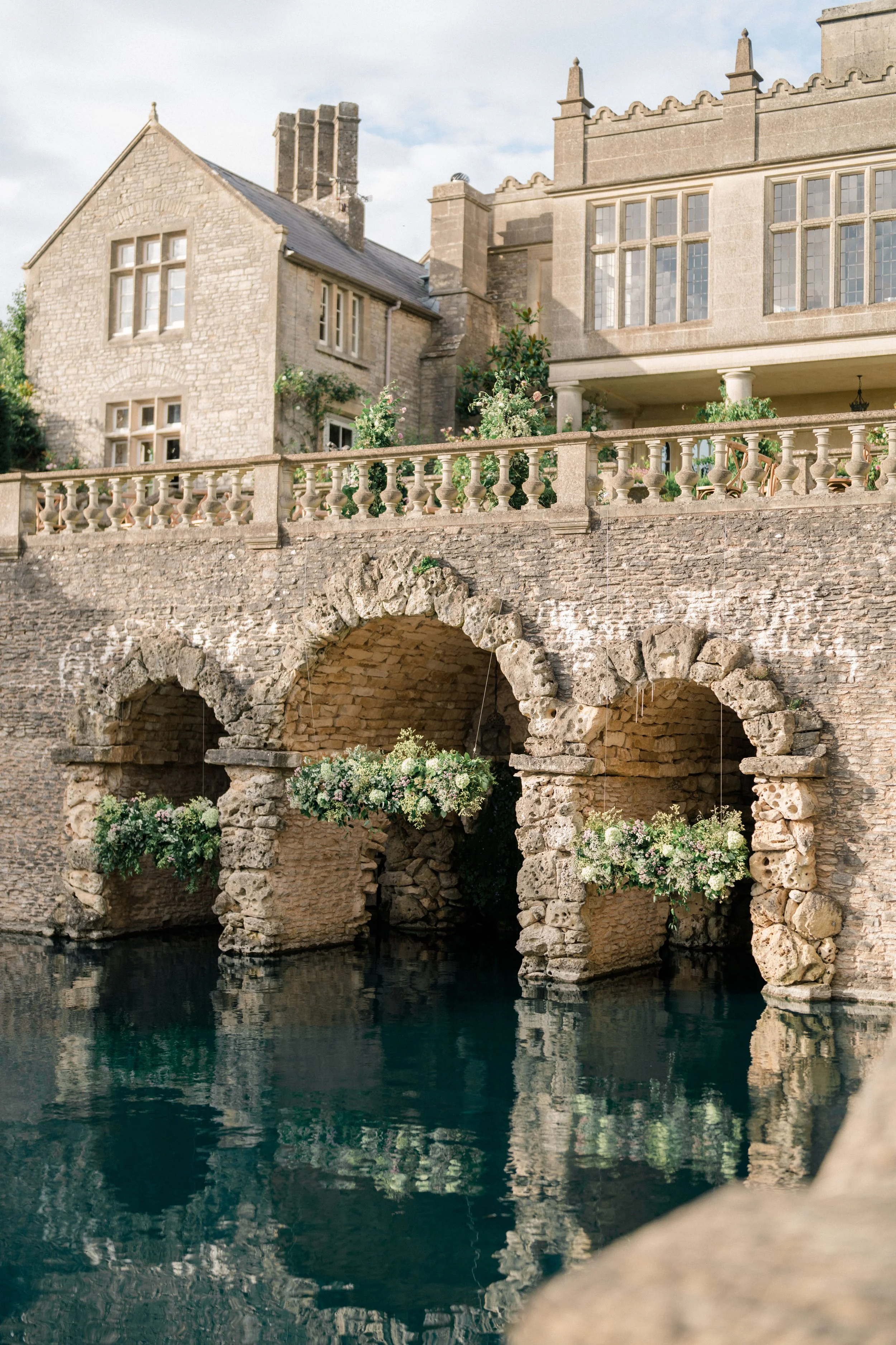 Euridge Manor Boat House with arches over water, adorned with greenery, under a clear sky in the Cotswolds, England, florist.
