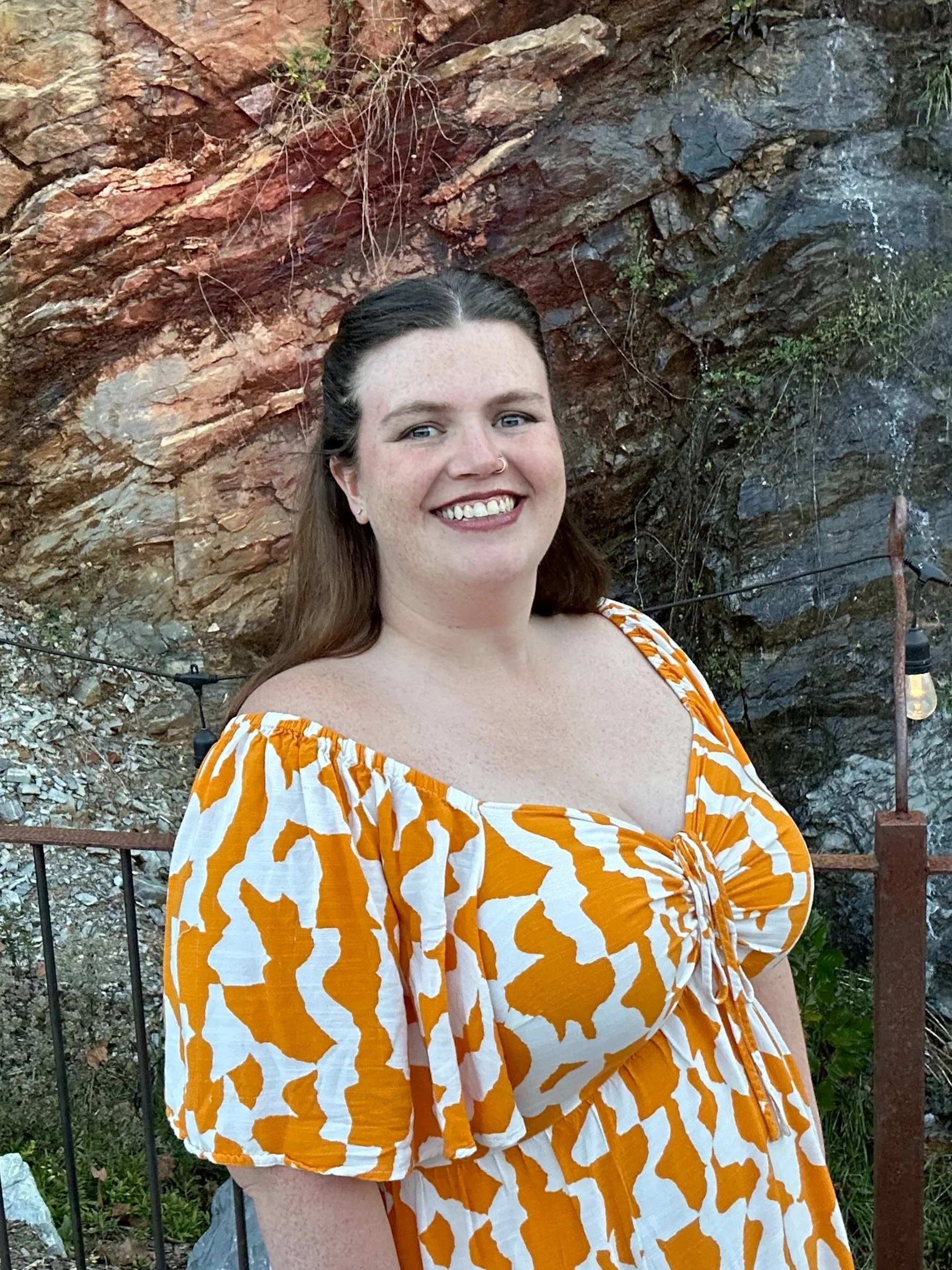 A woman smiling outdoors in front of a rocky background, wearing an orange and white patterned off-the-shoulder dress.