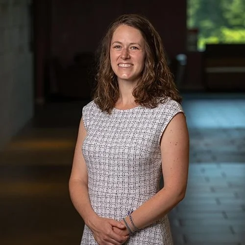 A woman with shoulder-length curly hair standing indoors, smiling, in a short-sleeved patterned dress.