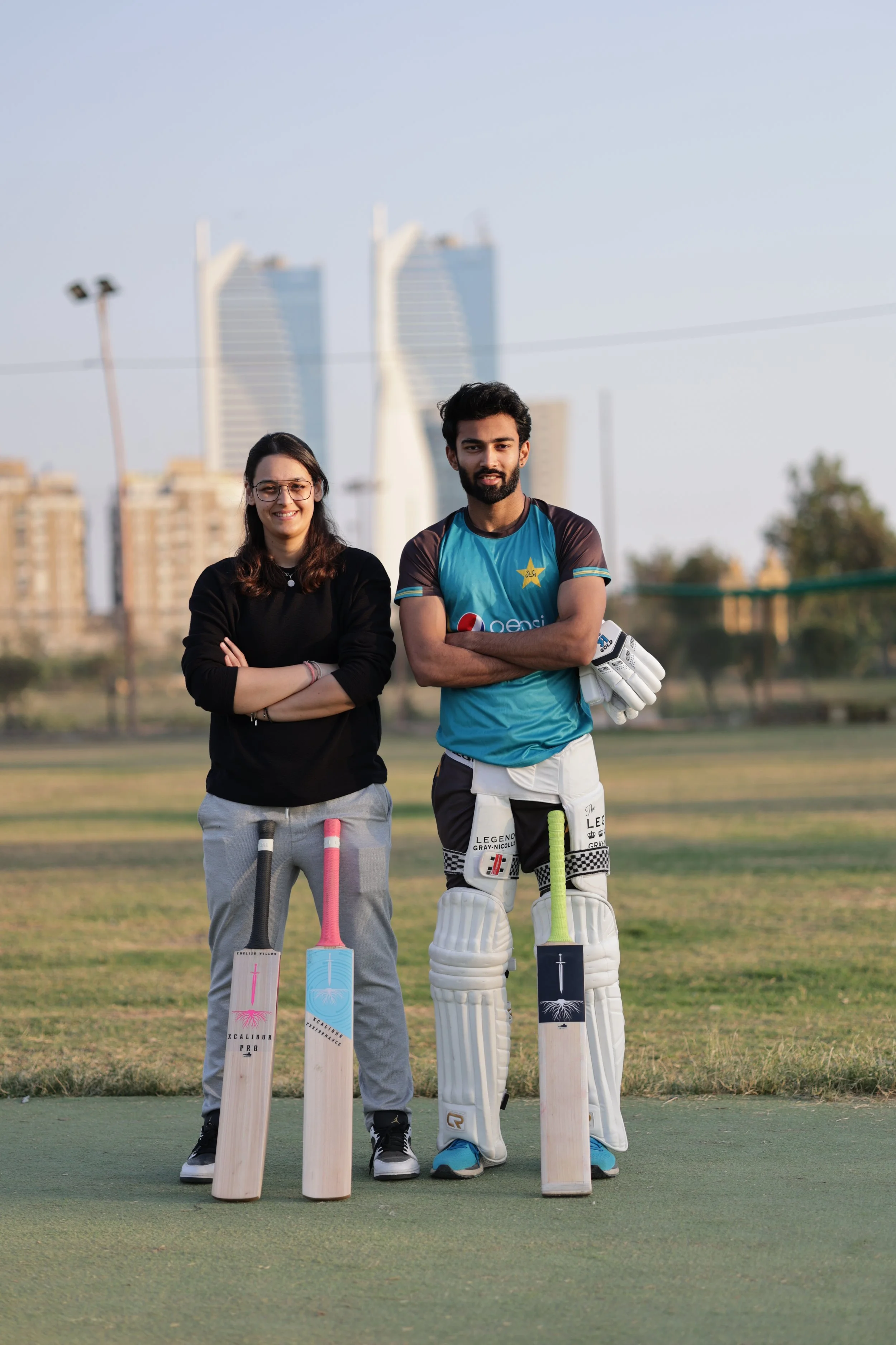 Two cricket players, a woman and a man, standing on a cricket ground with cricket bats. The woman has a black sweater and gray sweatpants, the man is in a blue cricket uniform with protective gear. In the background, there are tall buildings and a clear sky.