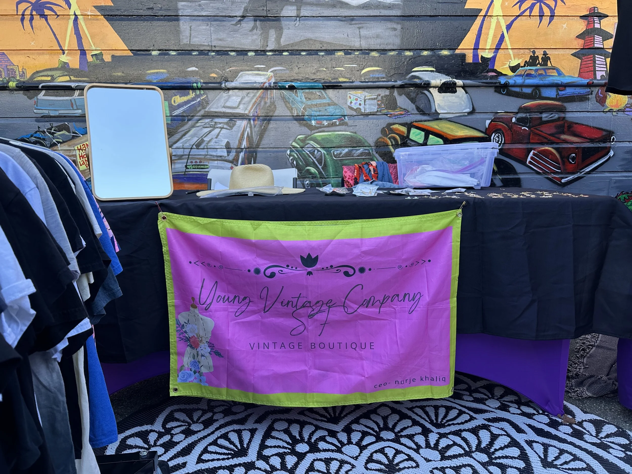 A colorful mural with cars, palm trees, and silhouetted people under a sunset sky. In front, there is a table with various clothing items and a sign that reads 'Young Vintage Company SF, Vintage Boutique'.
