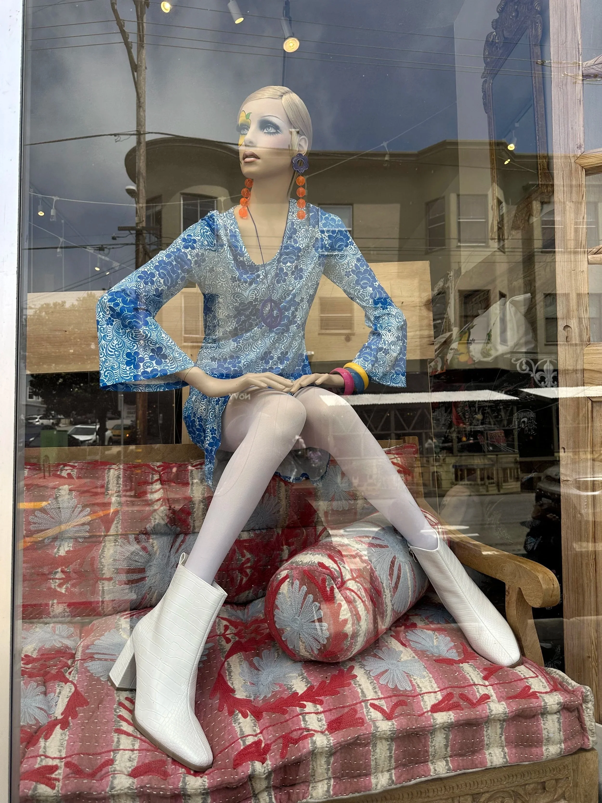 Twiggy Mannequin with blonde hair, wearing a blue floral patterned dress and large orange earrings, sitting on a sofa with floral upholstery, inside a store window, with reflections of the street outside.