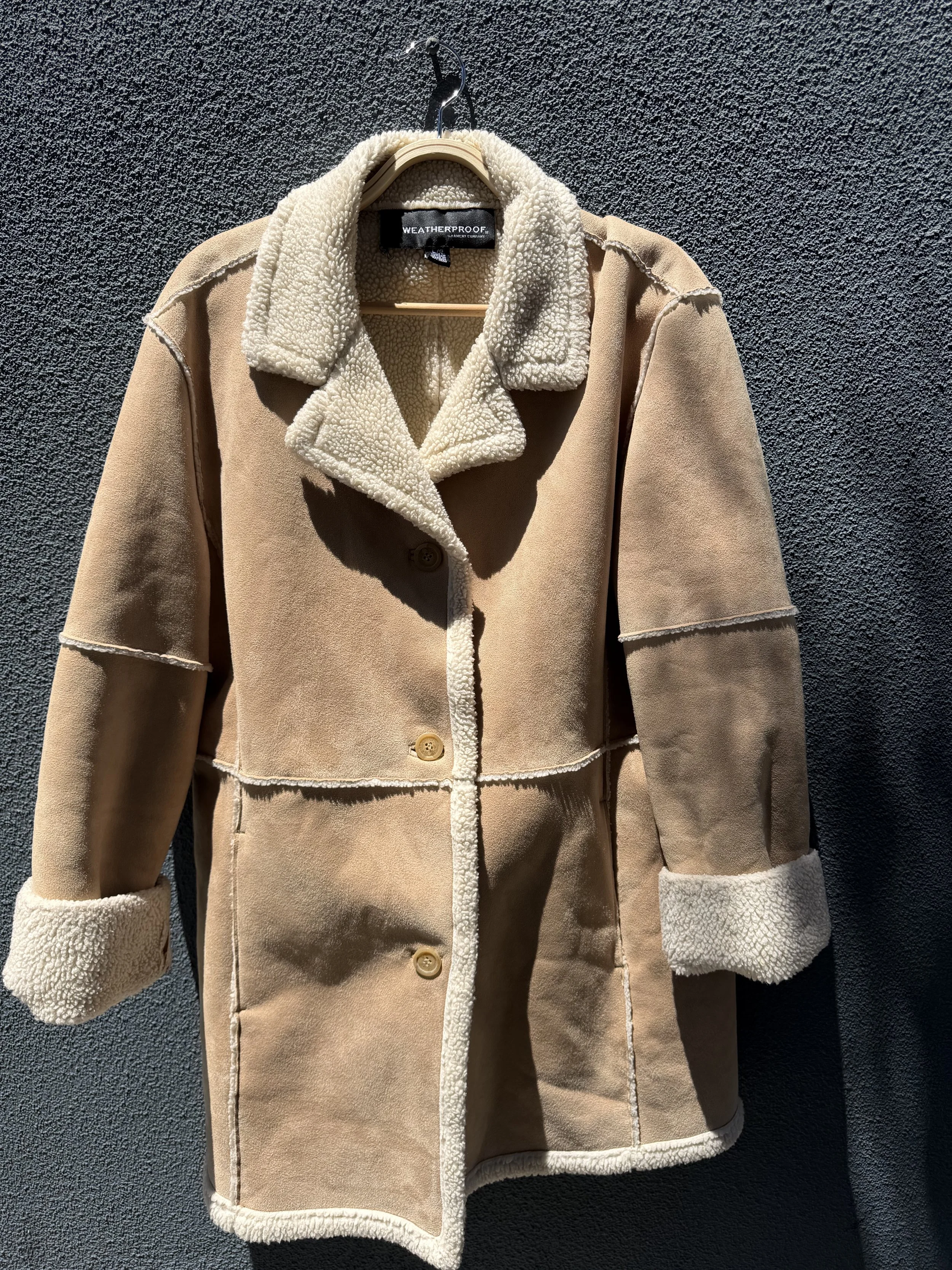A beige weatherproof coat with a faux shearling collar and cuffs, hanging on a black textured wall. The coat features a button closure.