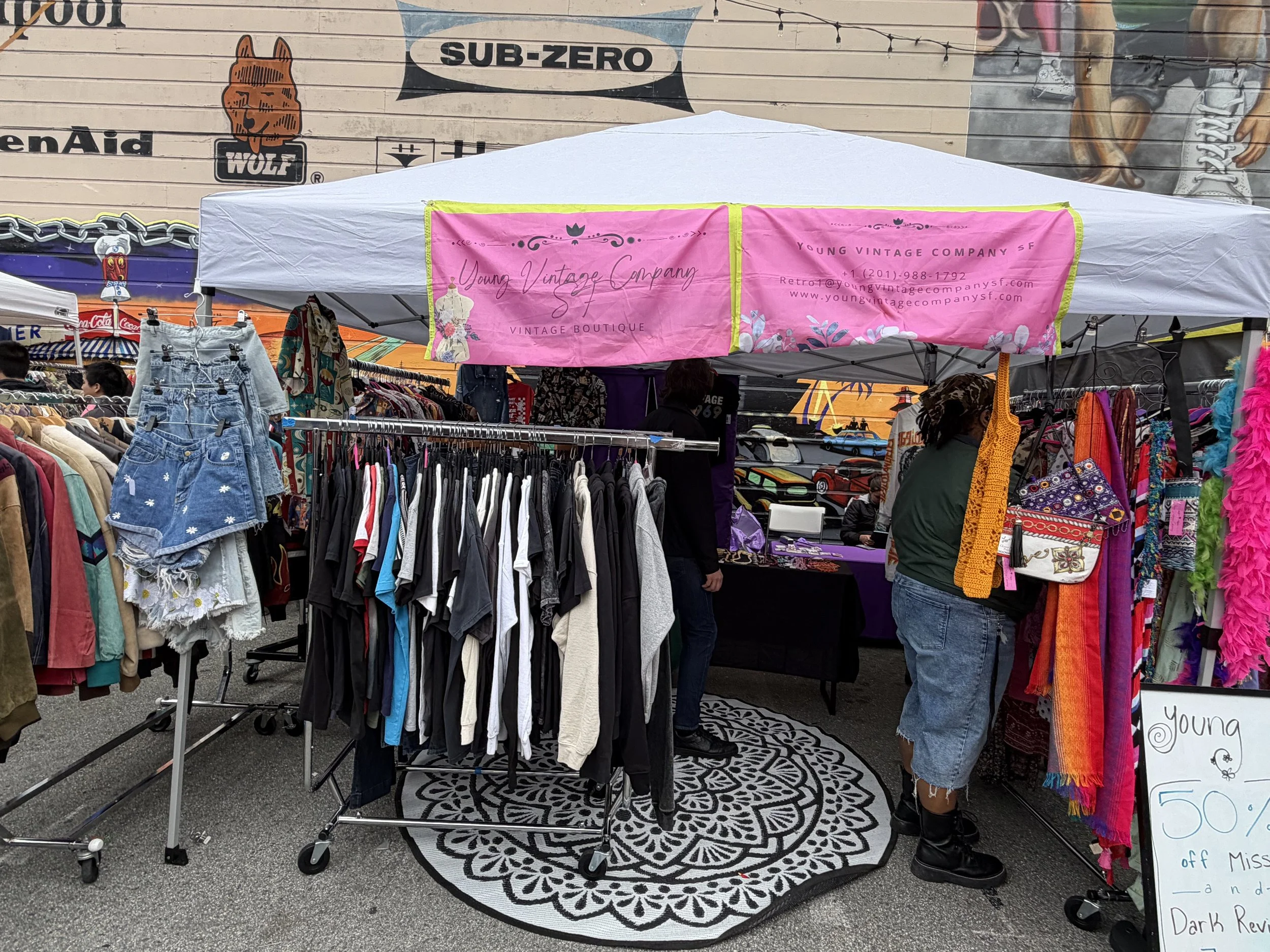 A clothing stall at an outdoor market with racks of clothes and accessories. A pink banner hangs above the stall, reading 'Young Vintage Company' and 'Vintage Boutique,' with contact info and website. Several people browse the items, and a mural with various artwork and characters is visible in the background.