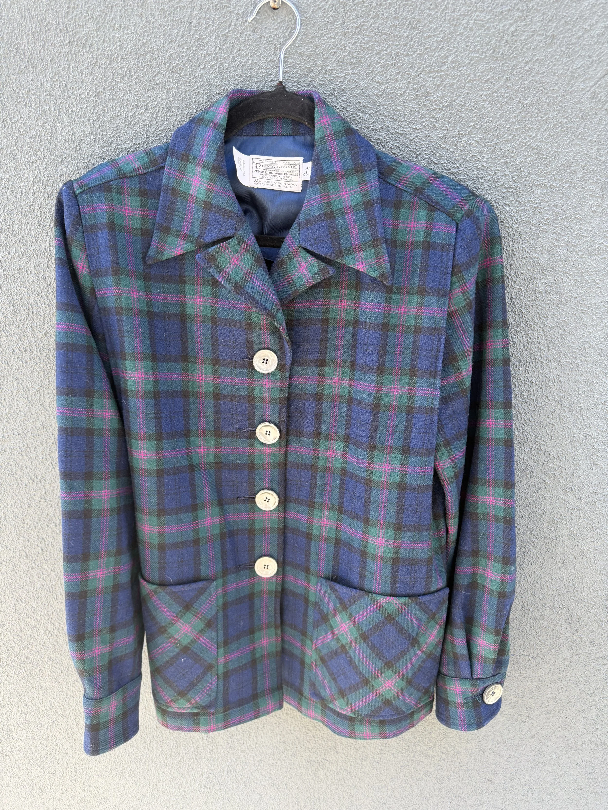 A plaid blazer in blue, green, and pink hangs on a black hanger against a gray textured wall.