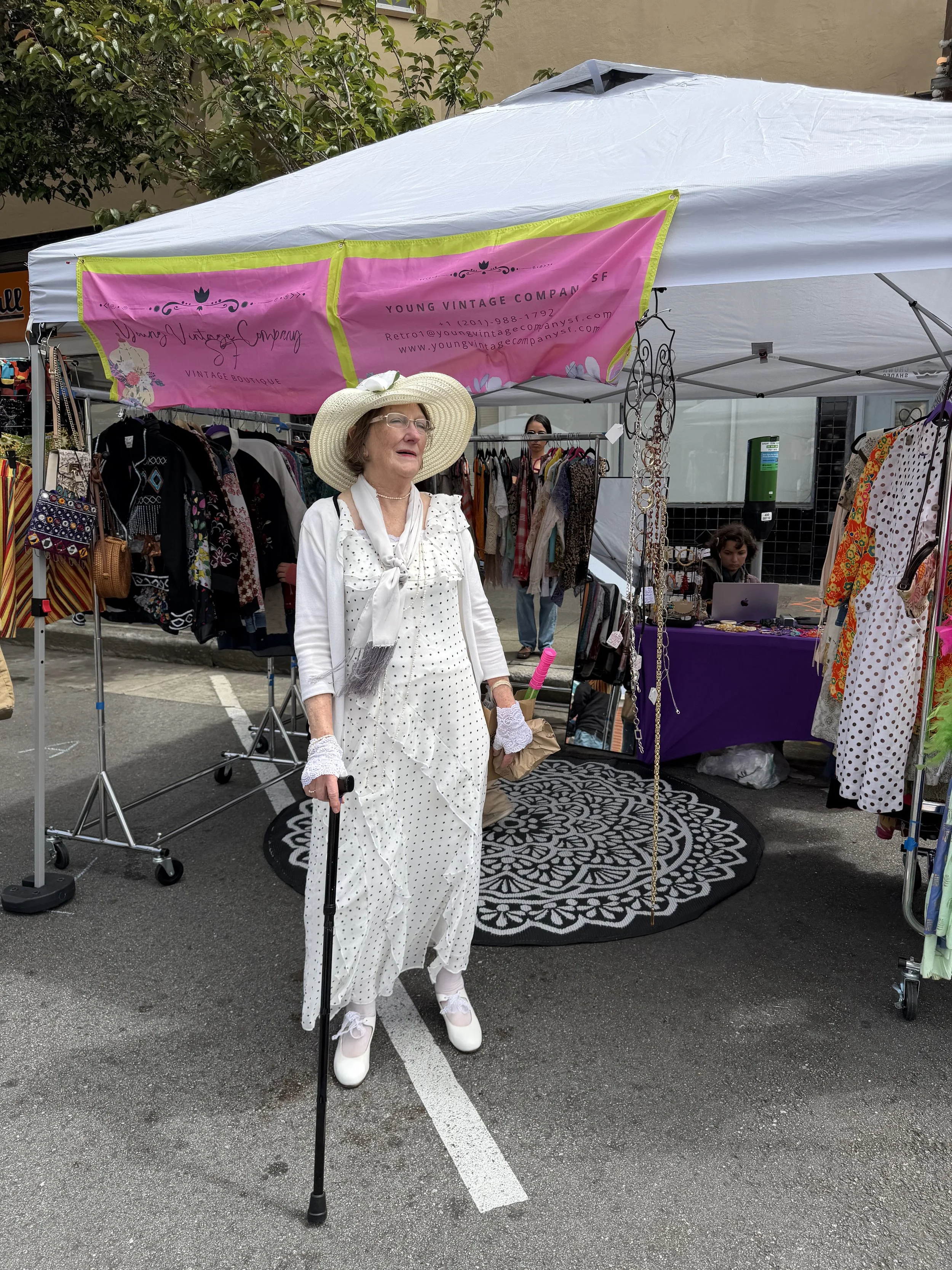 Woman dressed in vintage fashion standing under a pink and yellow vendor booth at an outdoor market. She is wearing a long white polka dot dress, a wide-brimmed hat with a bow, white gloves, and white shoes. Behind her are racks of clothing and jewelry, with other people visible in the background.