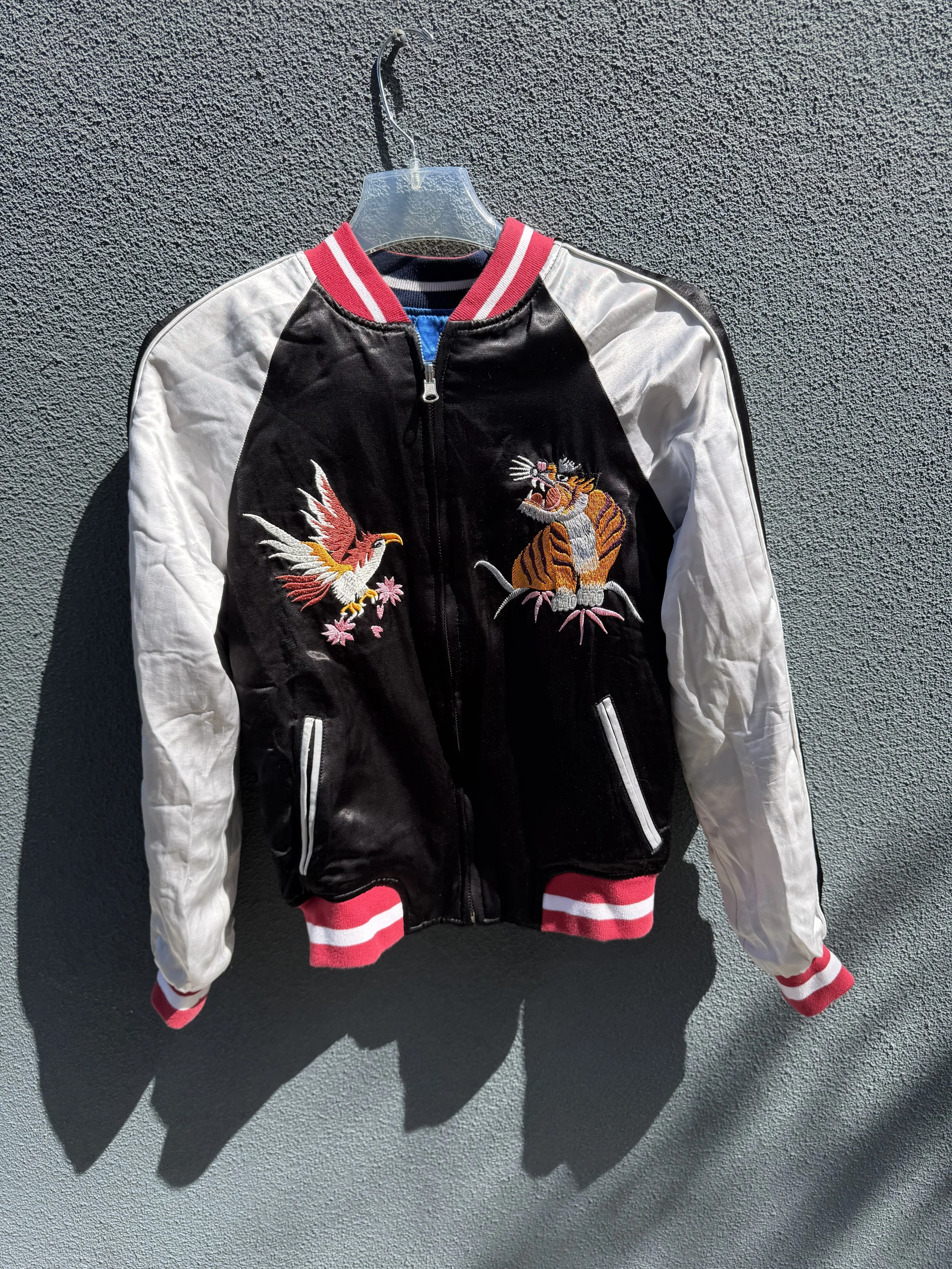 A black, white, and red varsity-style jacket hanging on a plastic hanger against a textured wall, featuring embroidered images of a bird and a tiger on the front.