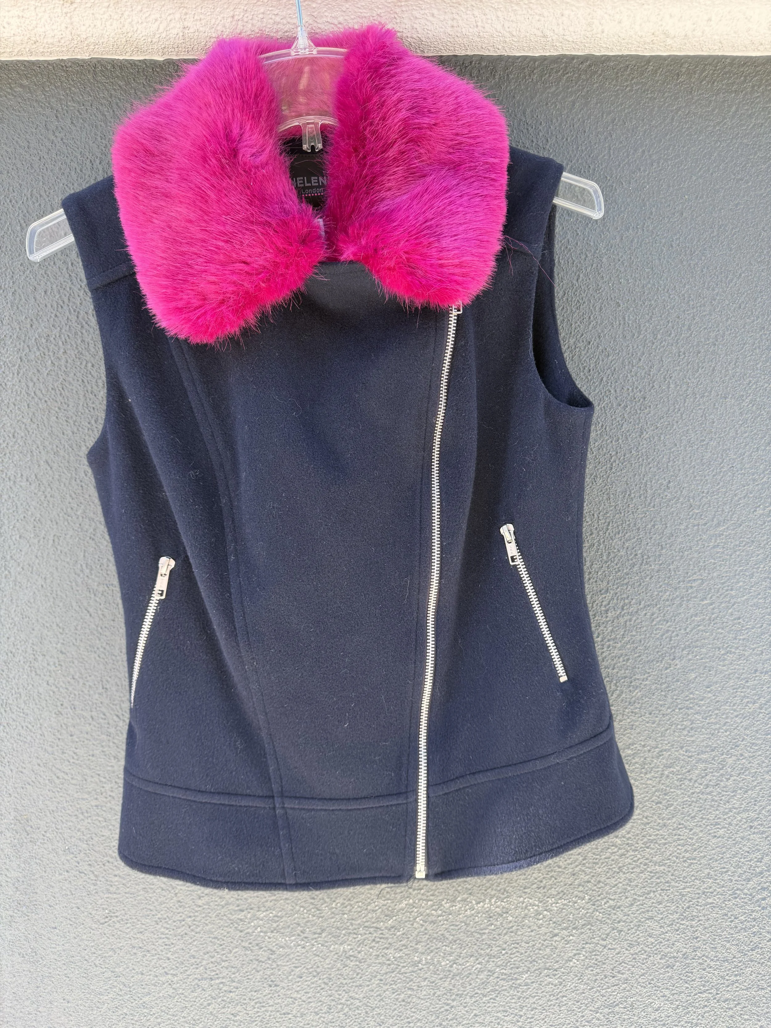 A sleeveless navy blue vest with silver zippers, hanging on a clear plastic hanger with bright pink faux fur around the collar, against a textured gray wall.