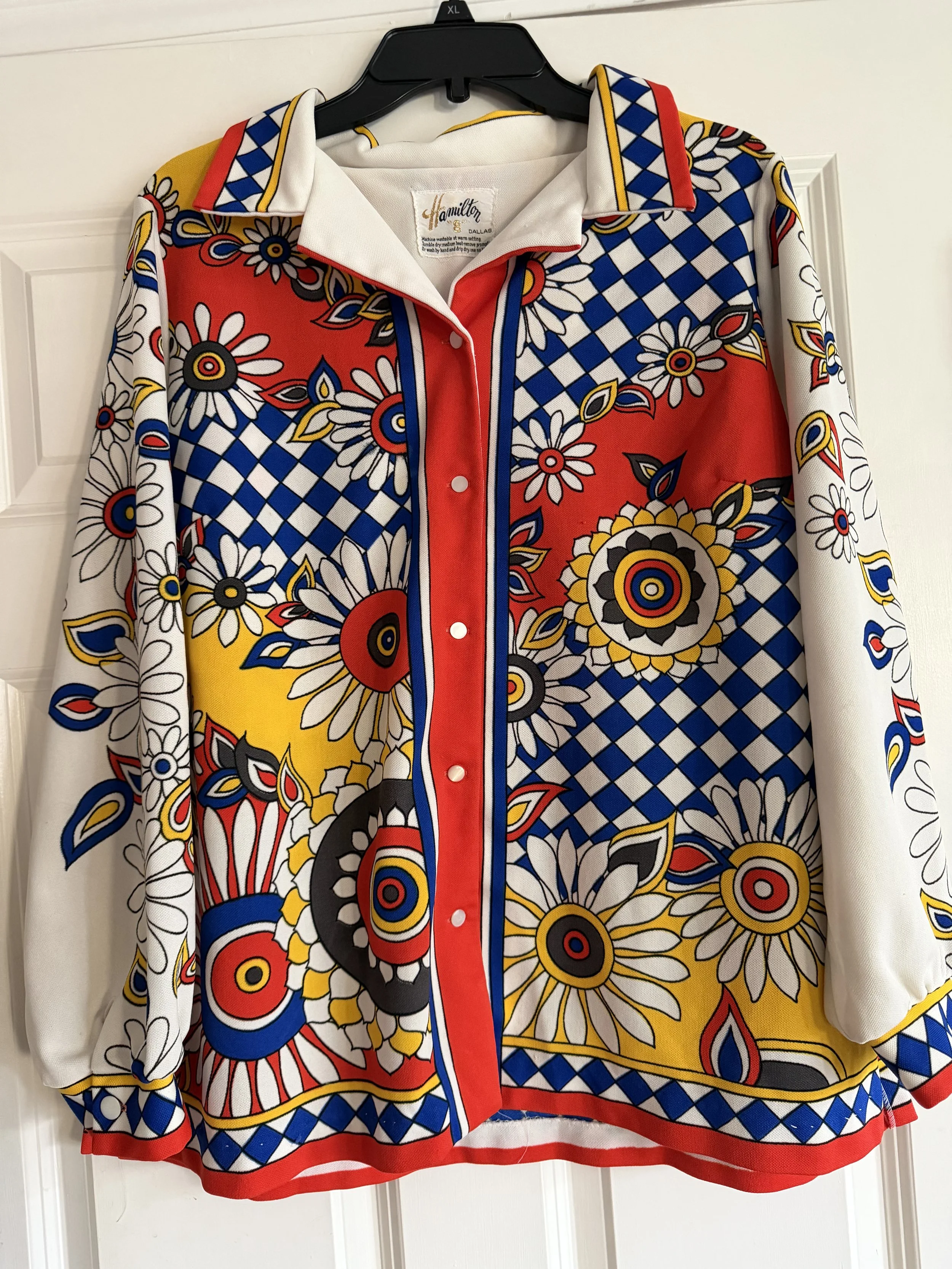 Colorful long-sleeve shirt with a floral and geometric pattern in red, yellow, blue, black, and white, hanging on a black hanger against a white door.