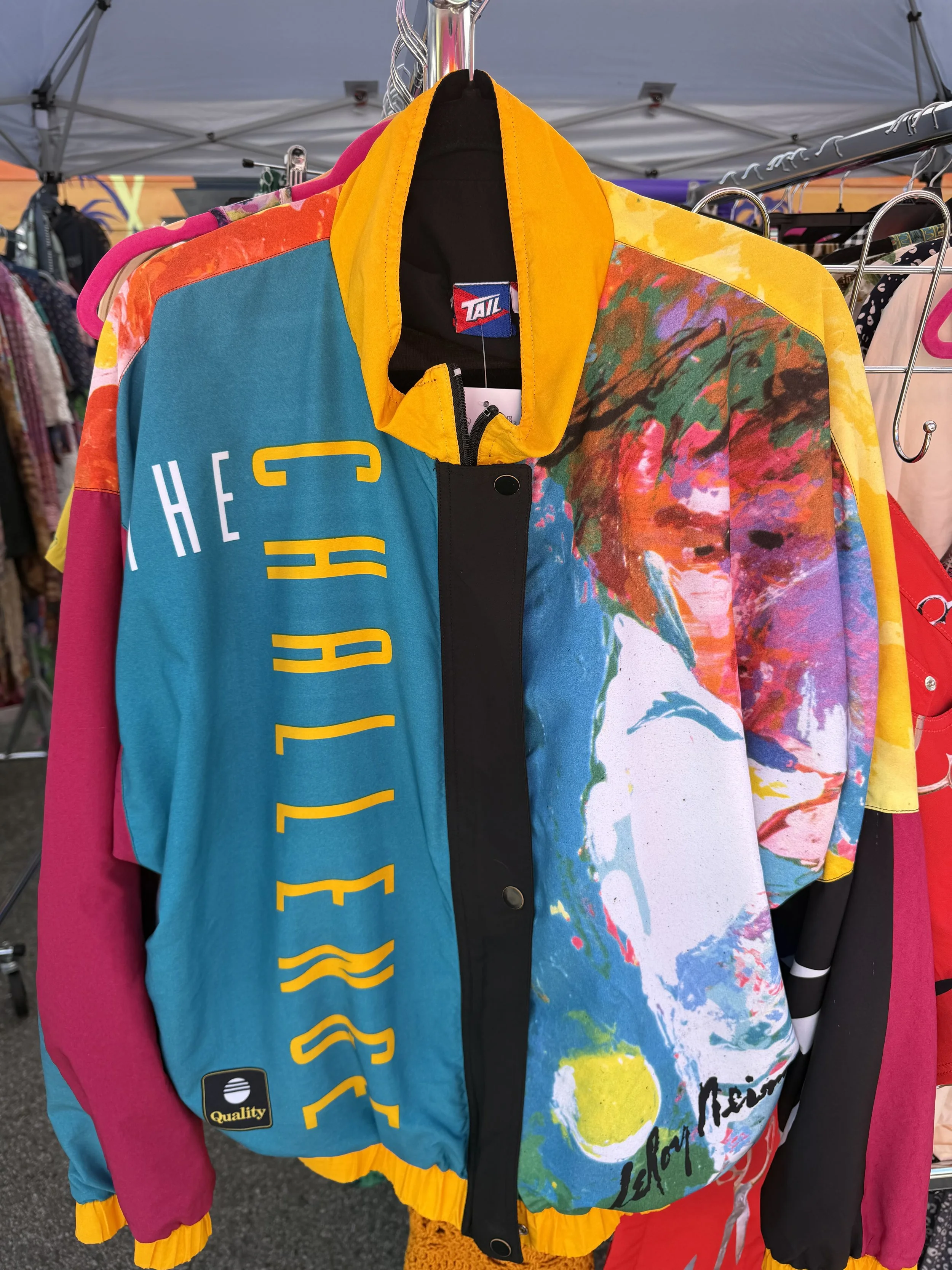 Colorful, abstract-patterned jackets hanging on a clothing rack at an outdoor market. The jackets feature bright colors including pink, blue, yellow, and multicolored designs.