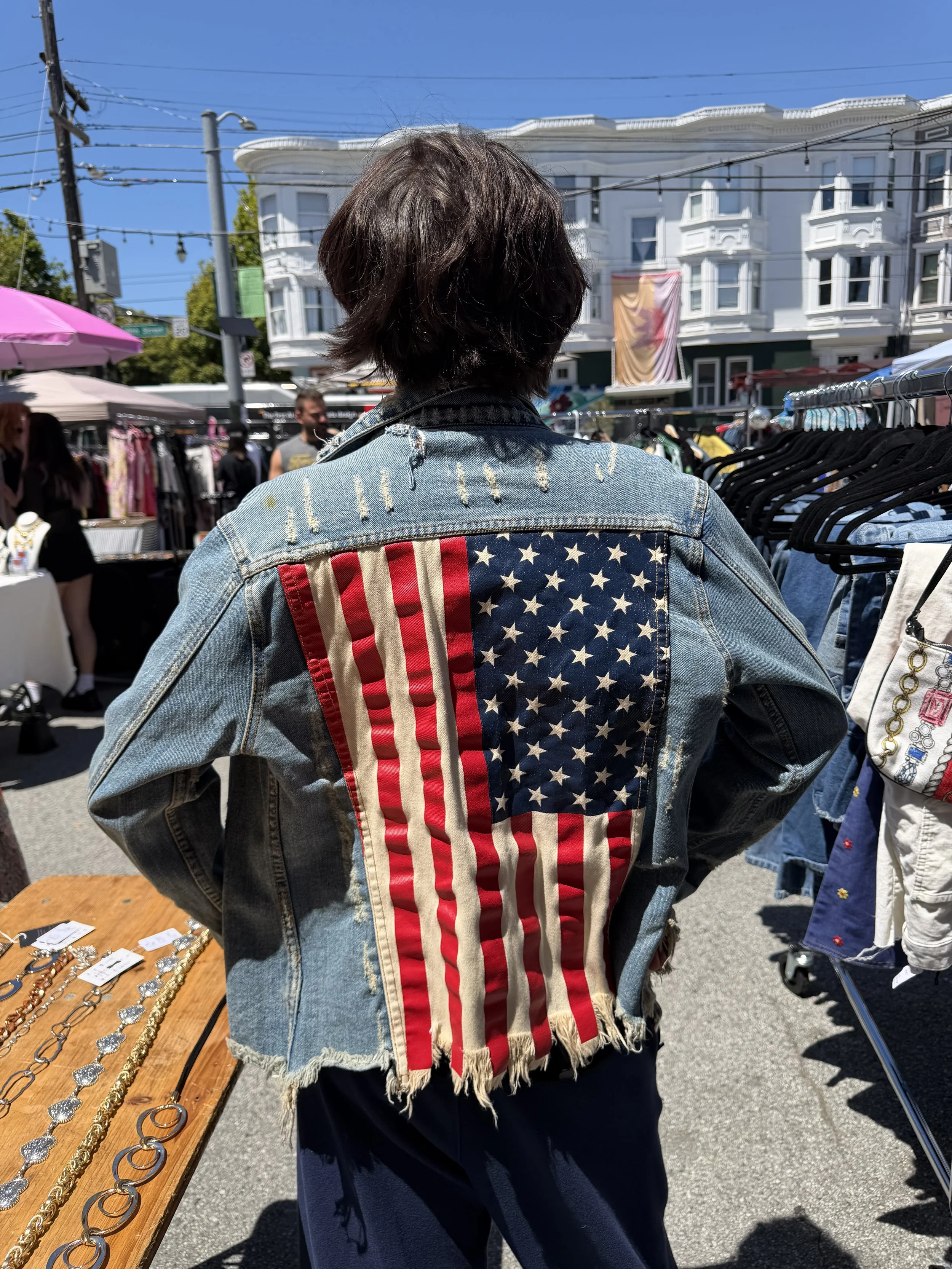Person standing at an outdoor market, wearing a denim jacket with an American flag design on the back, browsing through clothing racks under bright sunlight.