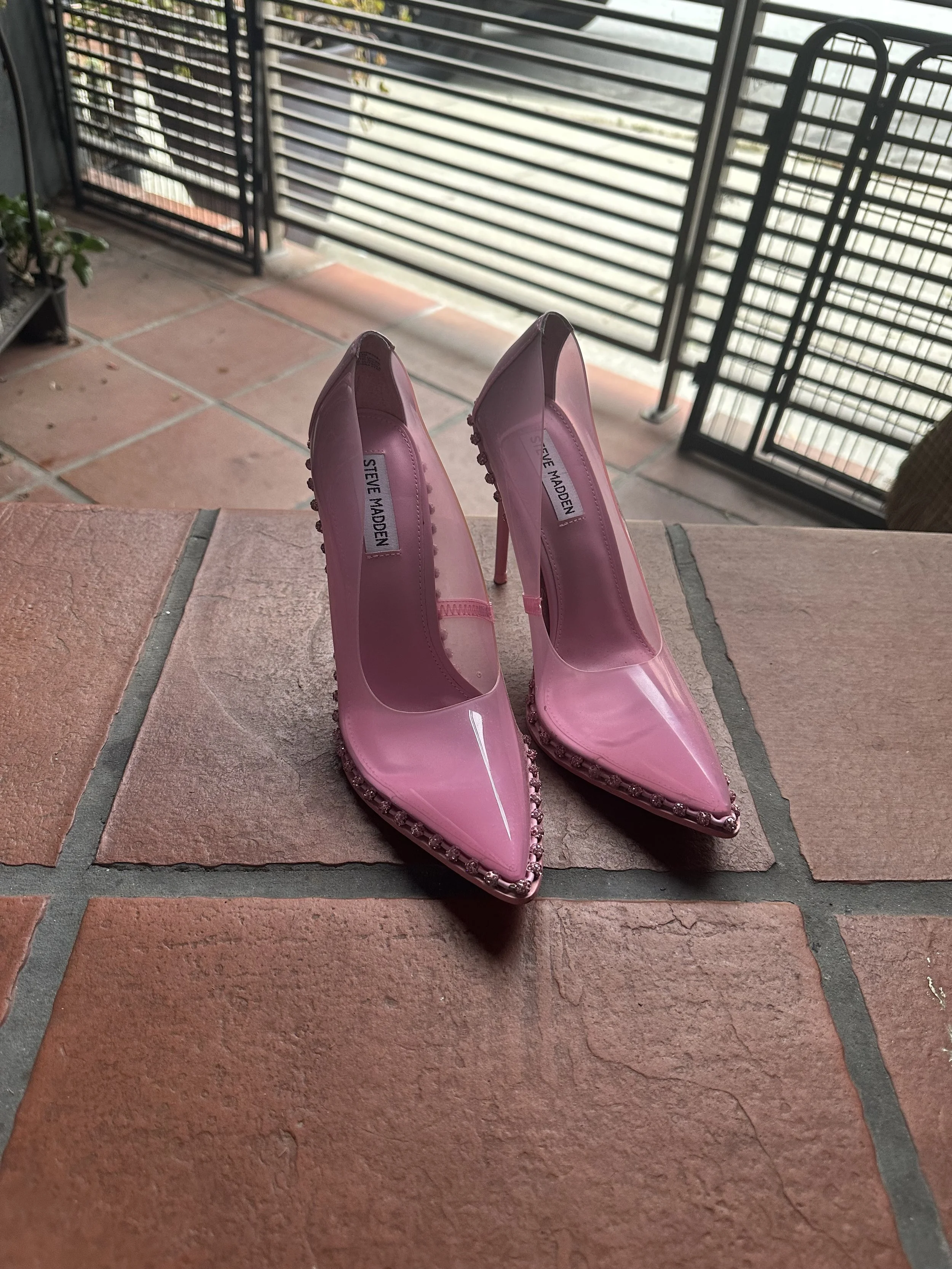 Pink pointed toe high heels with embellishments on the toe area, placed on a brick surface on a balcony or patio.
