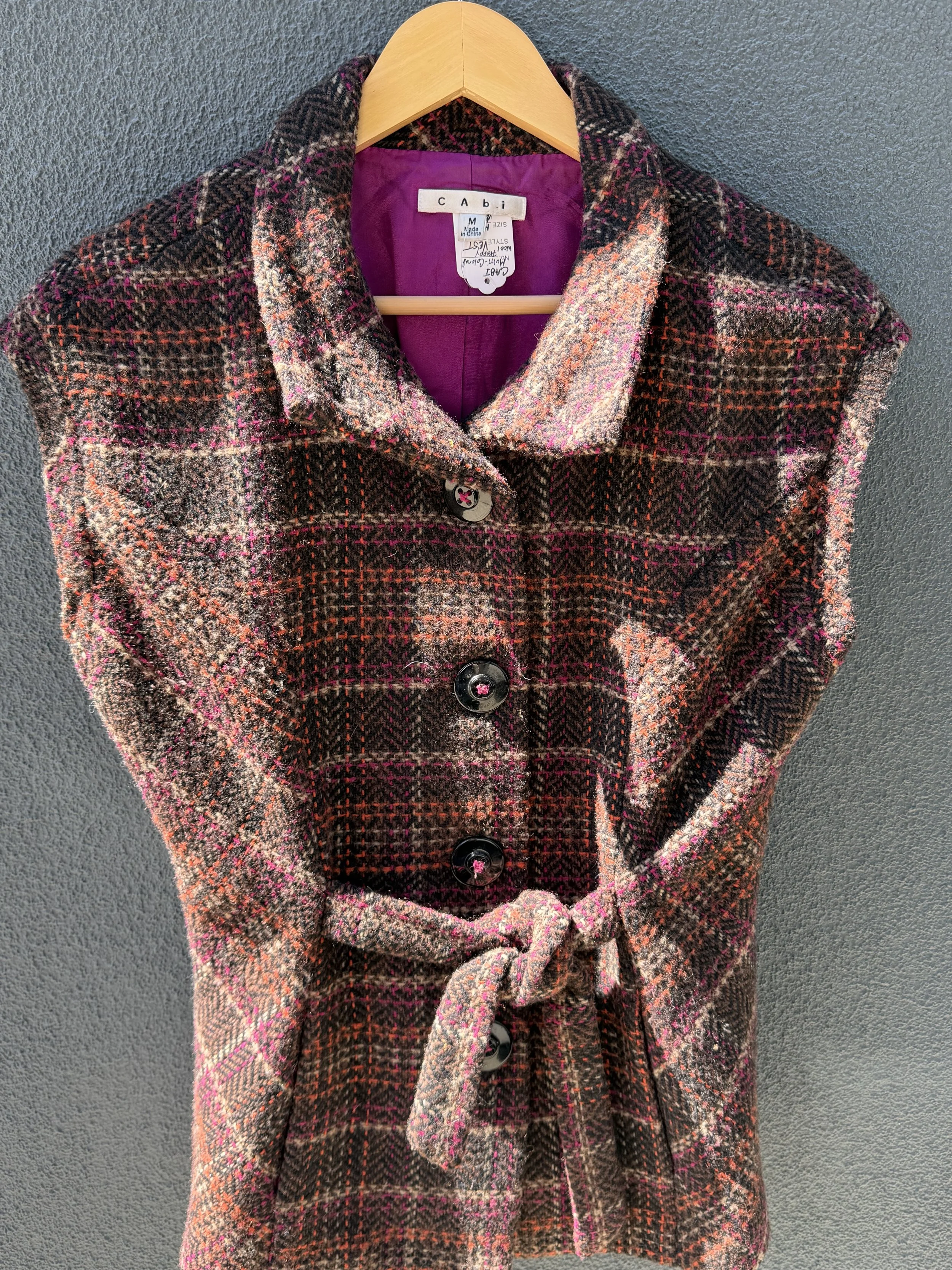 A plaid wool coat hanging on a wooden hanger against a gray wall. The coat has a mix of pink, black, brown, and cream colors, with large buttons down the front. The interior lining is purple.
