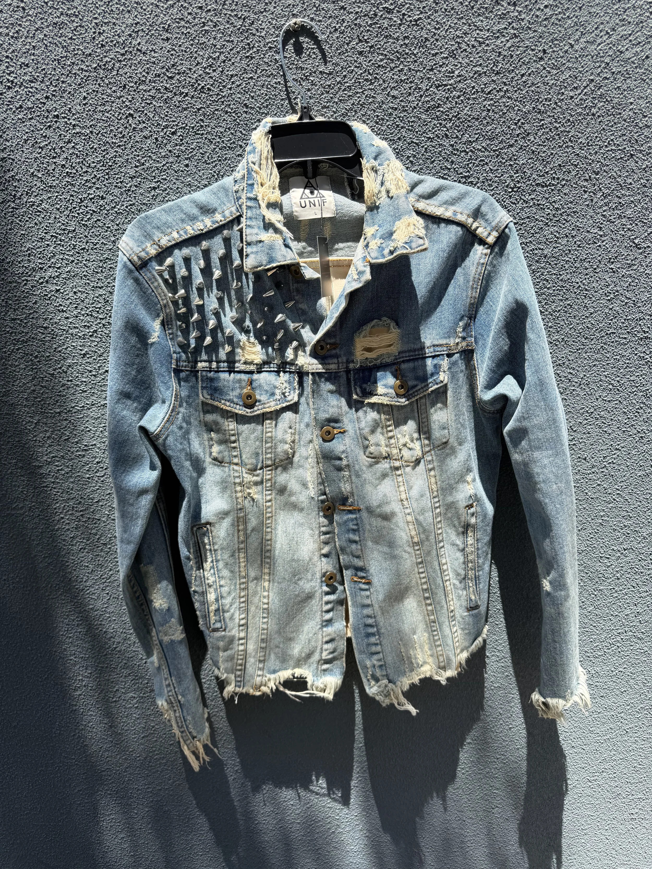 A distressed denim jacket with metal studs on the left shoulder and ripped fabric, hanging on a black hanger against a textured dark wall.