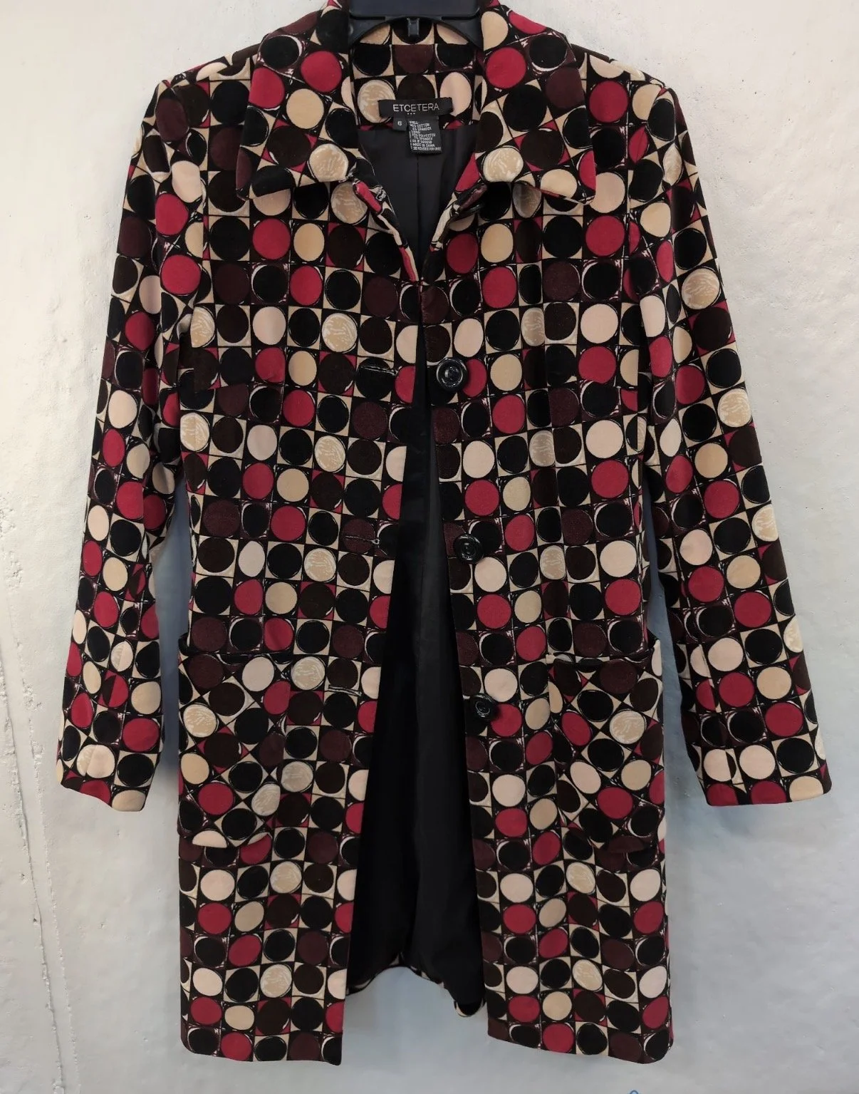 Long coat with a geometric pattern of circles in black, beige, red, and burgundy colors, hanging on a hanger against a plain white wall.