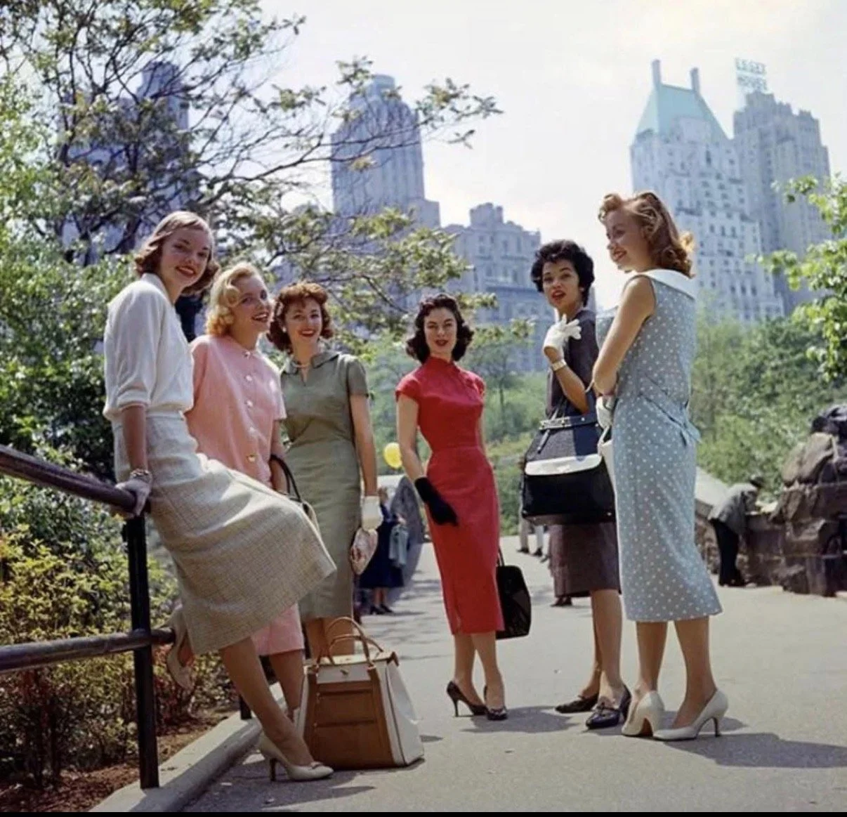 Seven women dressed in vintage 1950s-style clothing standing outdoors in a park, with skyscrapers in the background, smiling and chatting.