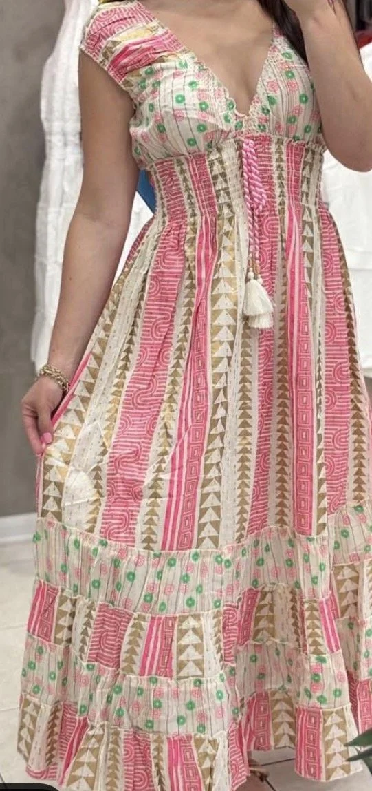 A woman wearing a colorful, bohemian-style dress with pink, green, beige, and white geometric and floral patterns. The dress has a v-neckline, cap sleeves, and a tiered, flowing skirt.