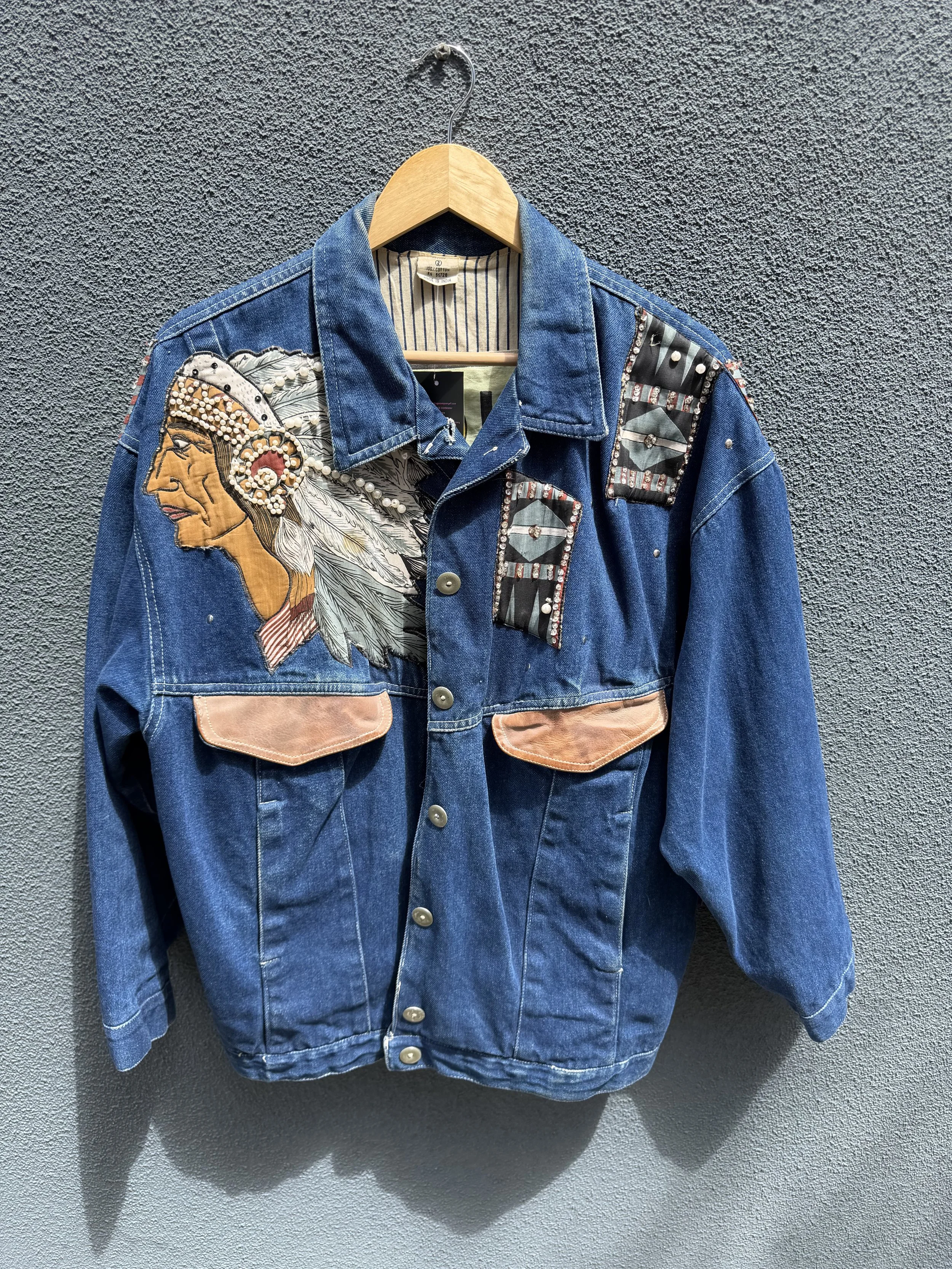 Blue denim jacket with embroidered and beaded Native American profile on the left chest, hanging on a wooden hanger against a textured gray wall.