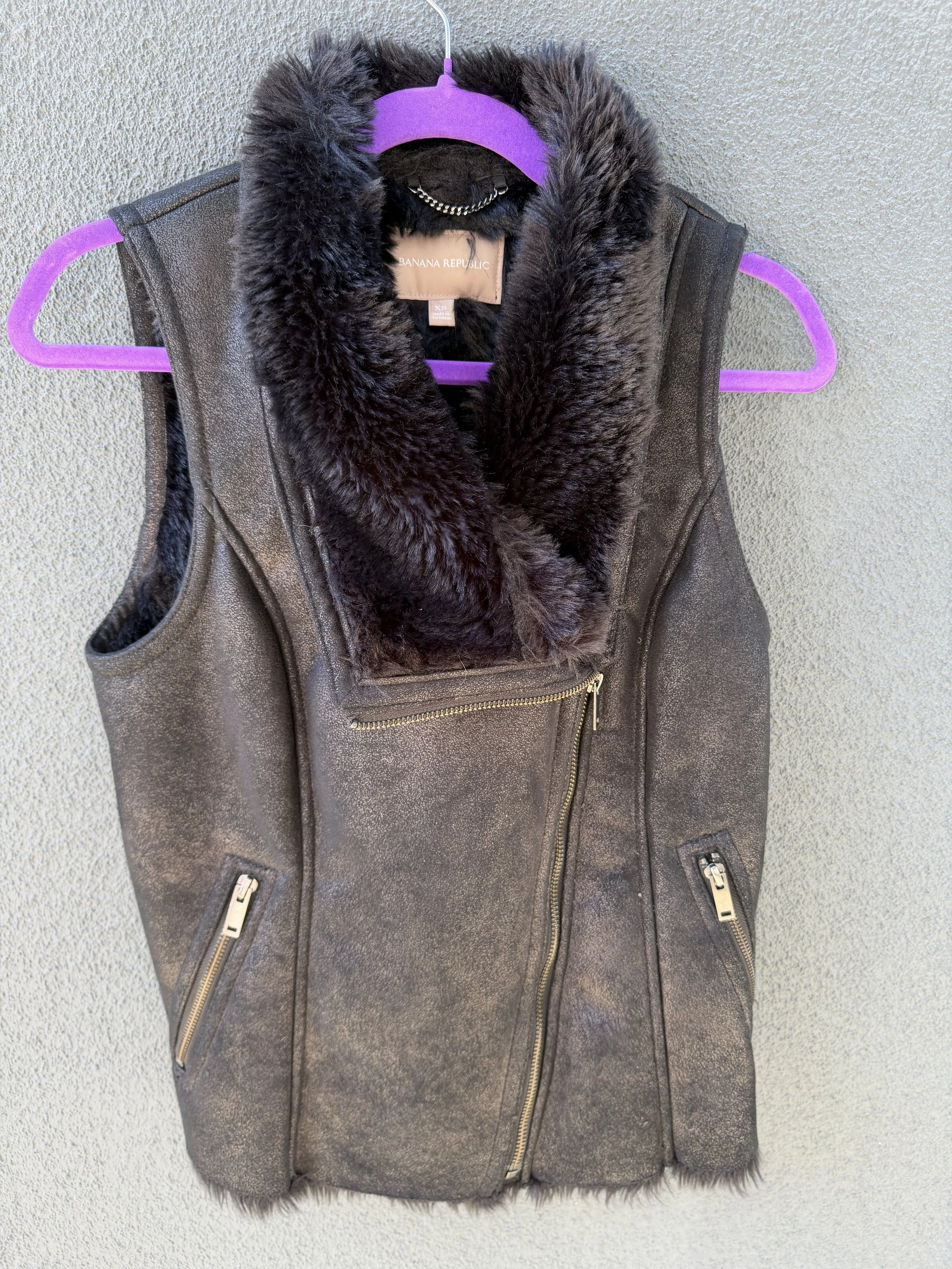 A dark brown sleeveless vest with gold zippers and a faux fur collar hanging on a purple hanger against a beige wall.