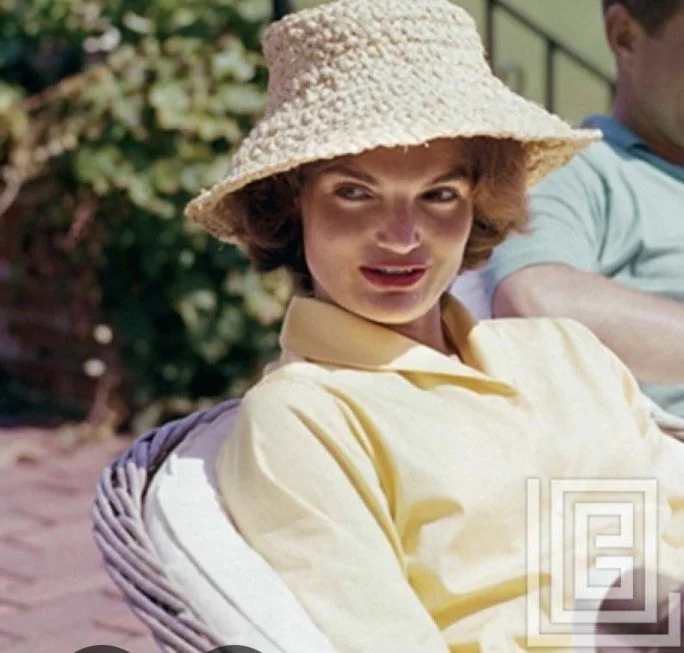 Woman wearing a wide-brimmed straw hat and a yellow shirt, sitting outdoors with blurred greenery in the background.