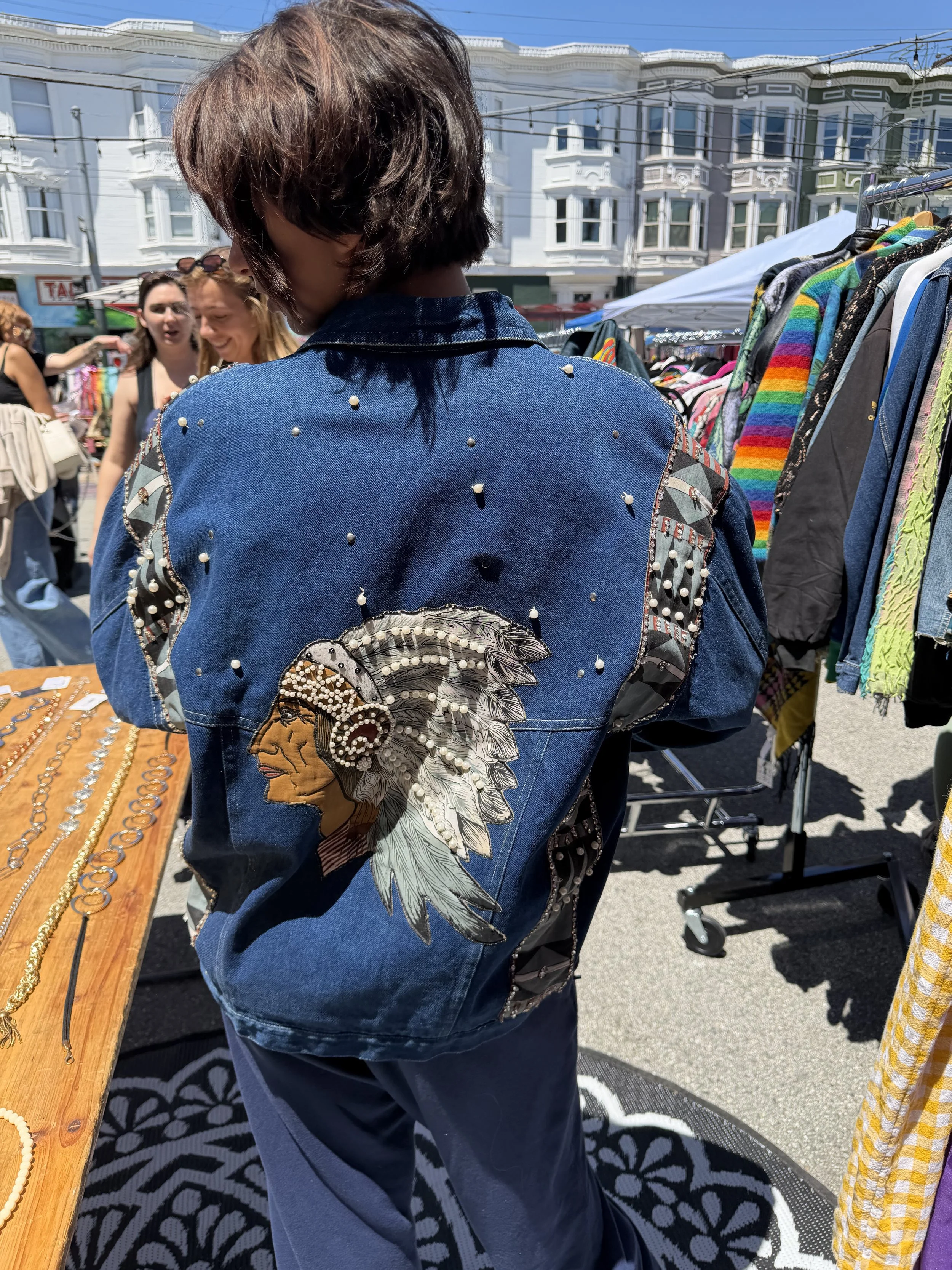 A person wearing a decorated blue denim jacket with pearl embellishments and a feathered patch, browsing outdoor market stalls with colorful clothing and jewelry, under a bright sunny sky.