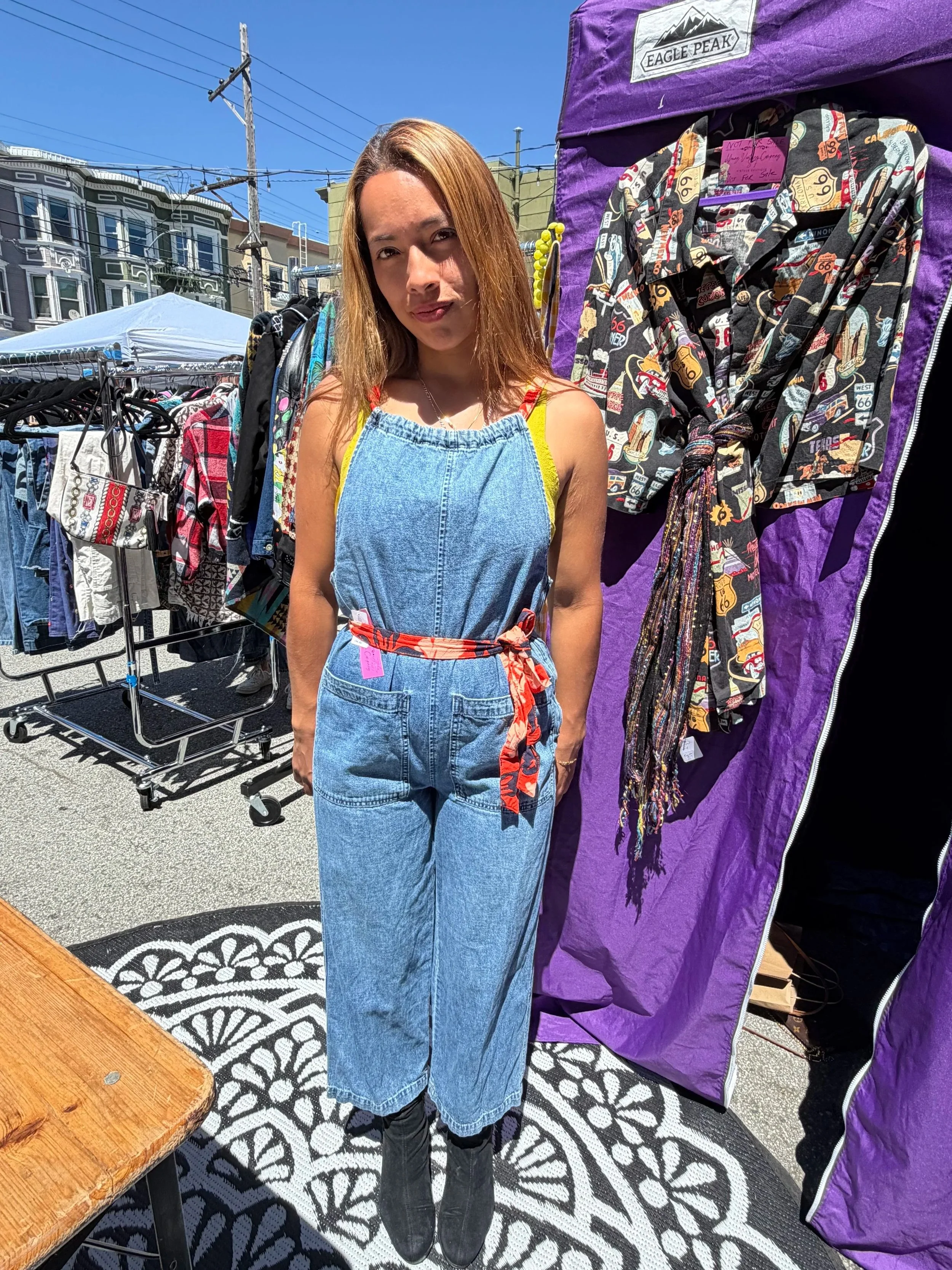 A young woman with long, light brown hair is standing outdoors in bright sunlight, wearing a denim dress with yellow straps and a colorful belt. She is standing next to a purple display tent with clothing on hangers. There are racks of clothes behind her and a sidewalk with a decorative black and white patterned mat.