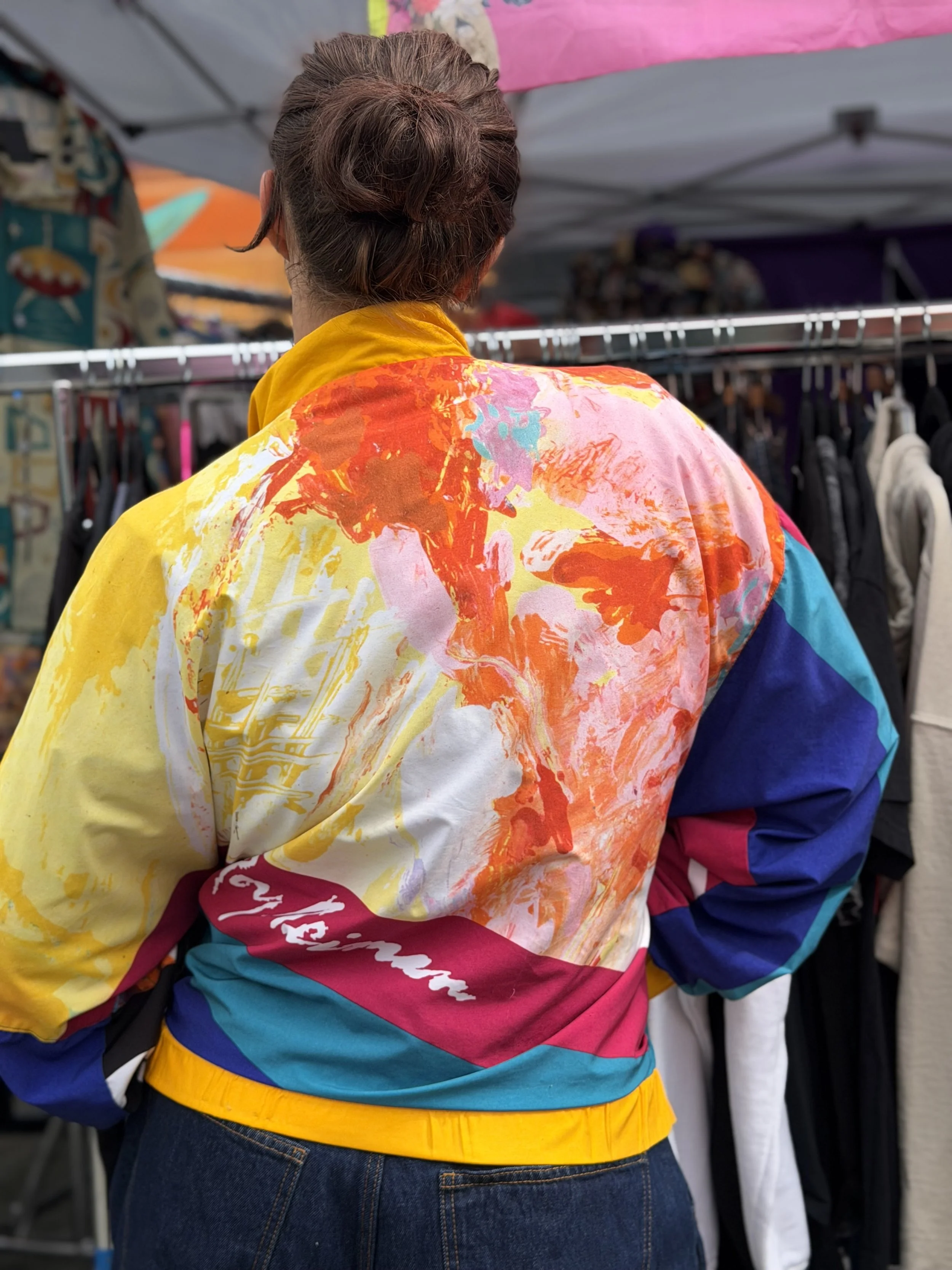 A person wearing a colorful, tie-dye style jacket with yellow, pink, purple, blue, and white hues, standing in front of a clothing rack with various garments.