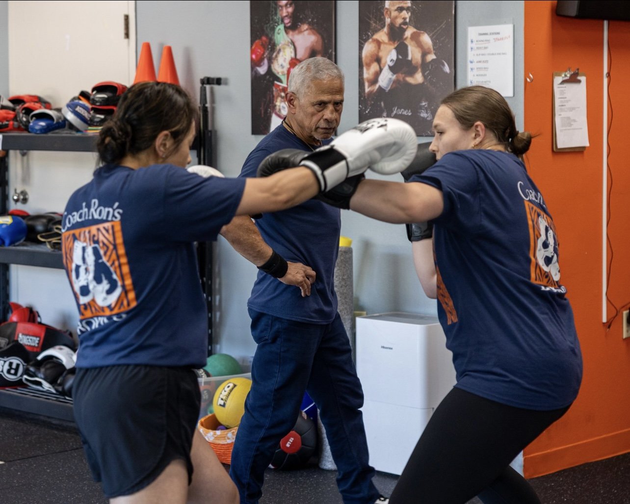 Two women at Coach Ron's Boxing practicing a punching drill while Coach Ron observers.